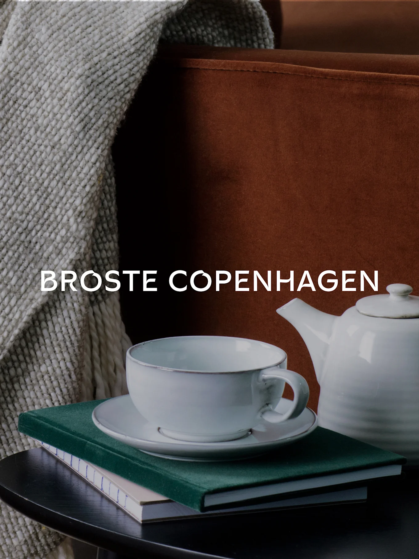 BROSTE COPENHAGEN text, a white teacup, teapot, and books on a table with a knitted blanket and sofa.