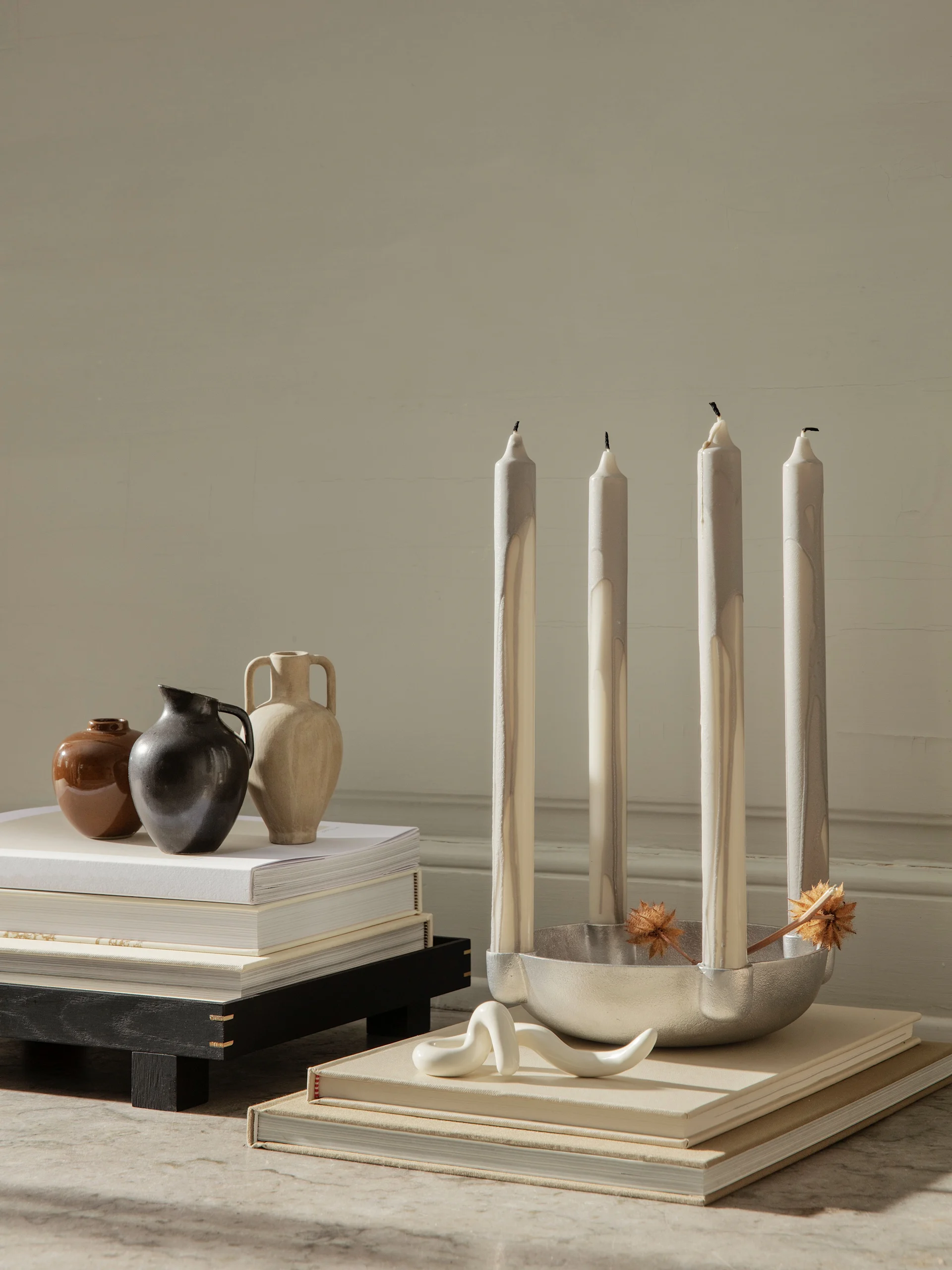Four tall, streaked candles in a silver holder with dried botanicals, a wavy white sculpture, three vases, and books.