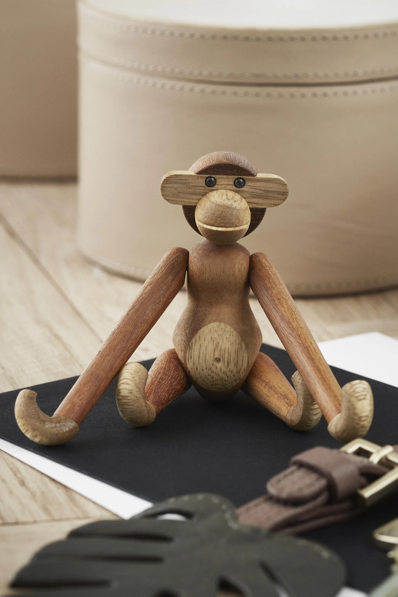 A wooden monkey figurine with articulated limbs sitting on a black surface, with blurred leather accessories and storage boxes in the background.