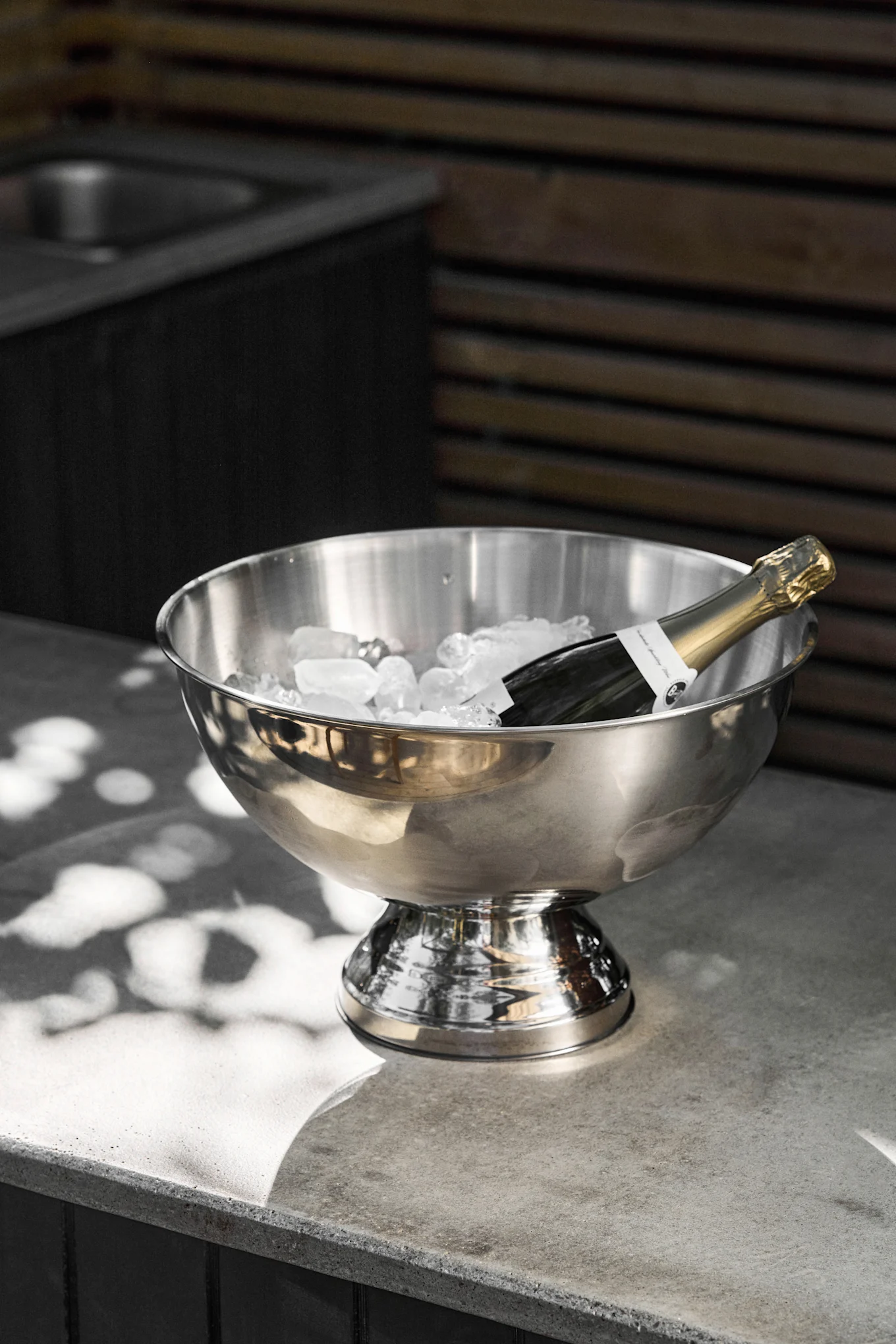 A champagne bottle rests in a stainless steel ice bucket filled with ice cubes on a gray concrete counter, with dappled sunlight.