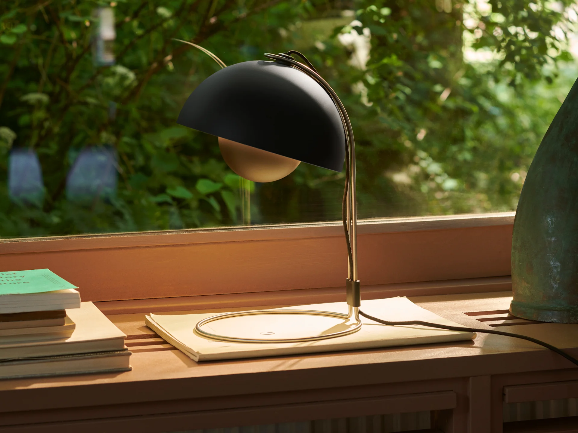 A black spherical desk lamp with a gold curved stand sits on a wooden shelf next to books, with green foliage visible outside a window.