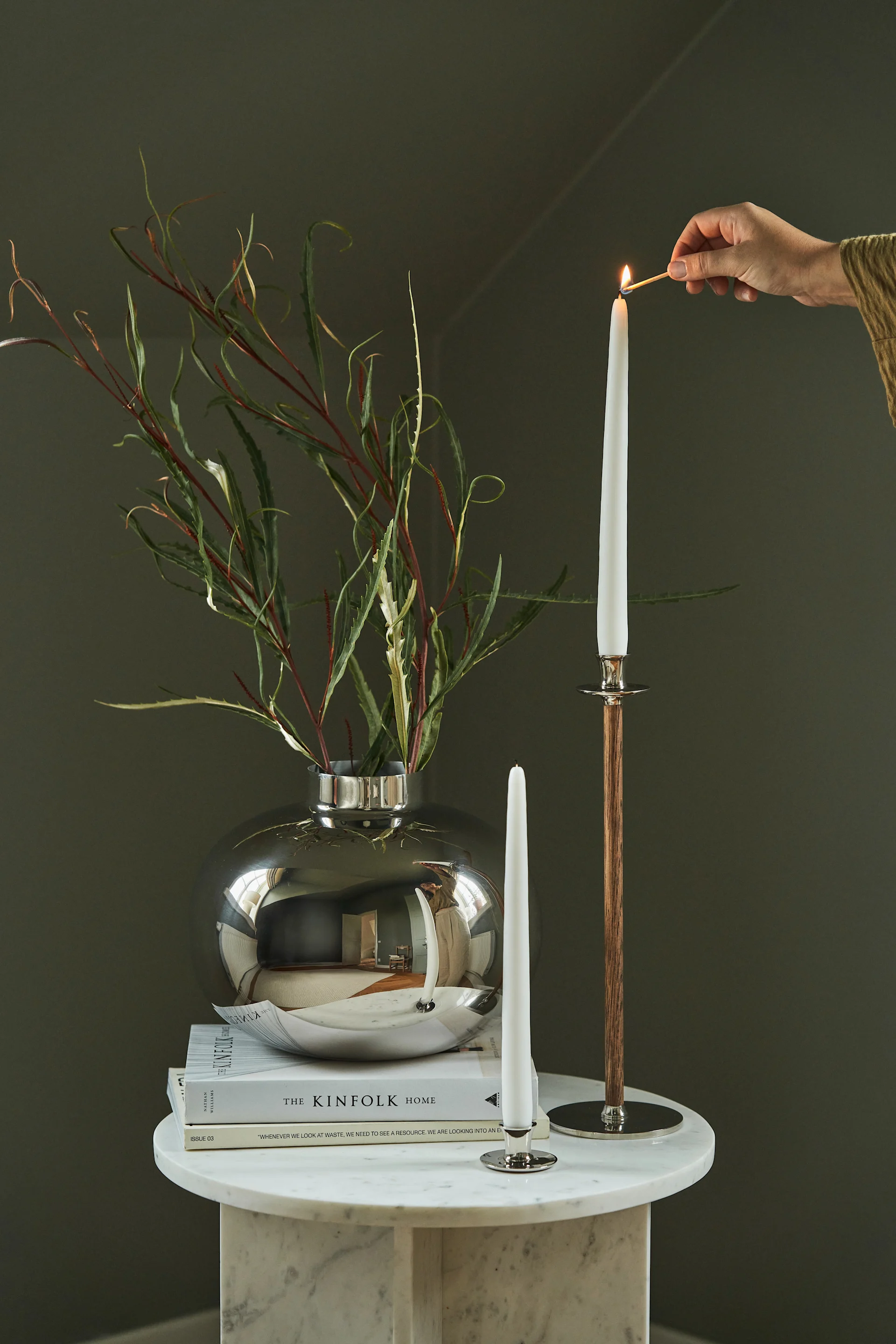 A hand lights a tall white candle on a wooden candelabra next to a silver vase with foliage and books on a marble table.