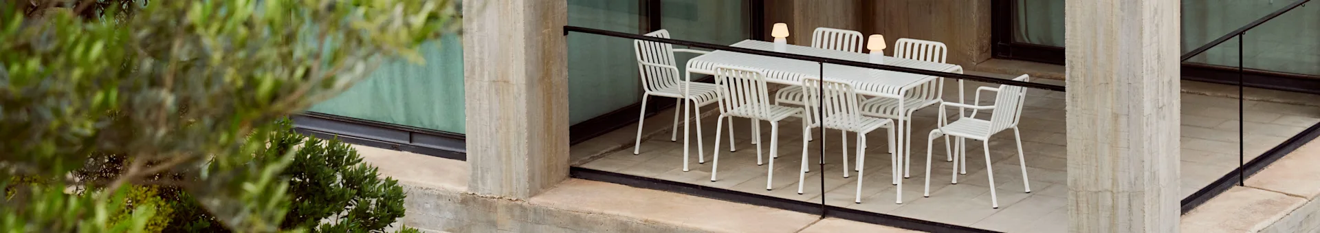 An outdoor dining set with a white slatted table and six matching chairs, lit by three portable lamps, on a concrete patio with plants.