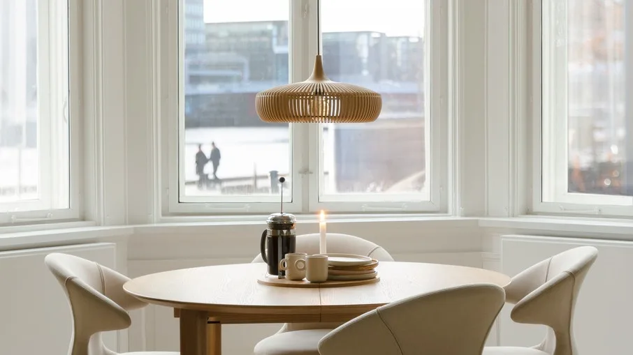 Round wooden dining table with four light chairs, a wooden slatted pendant lamp, French press, mugs, and plates on top.