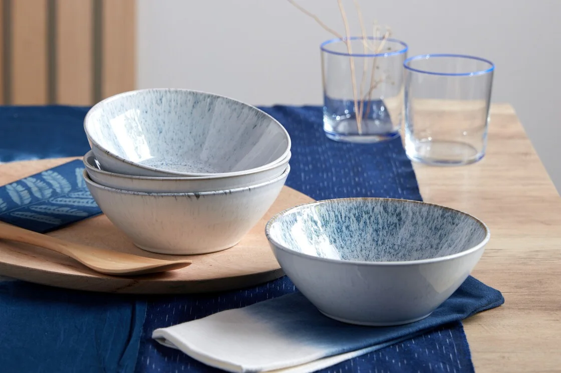 Table setting with blue speckled bowls, a wooden spoon, and clear glasses on a blue table runner.