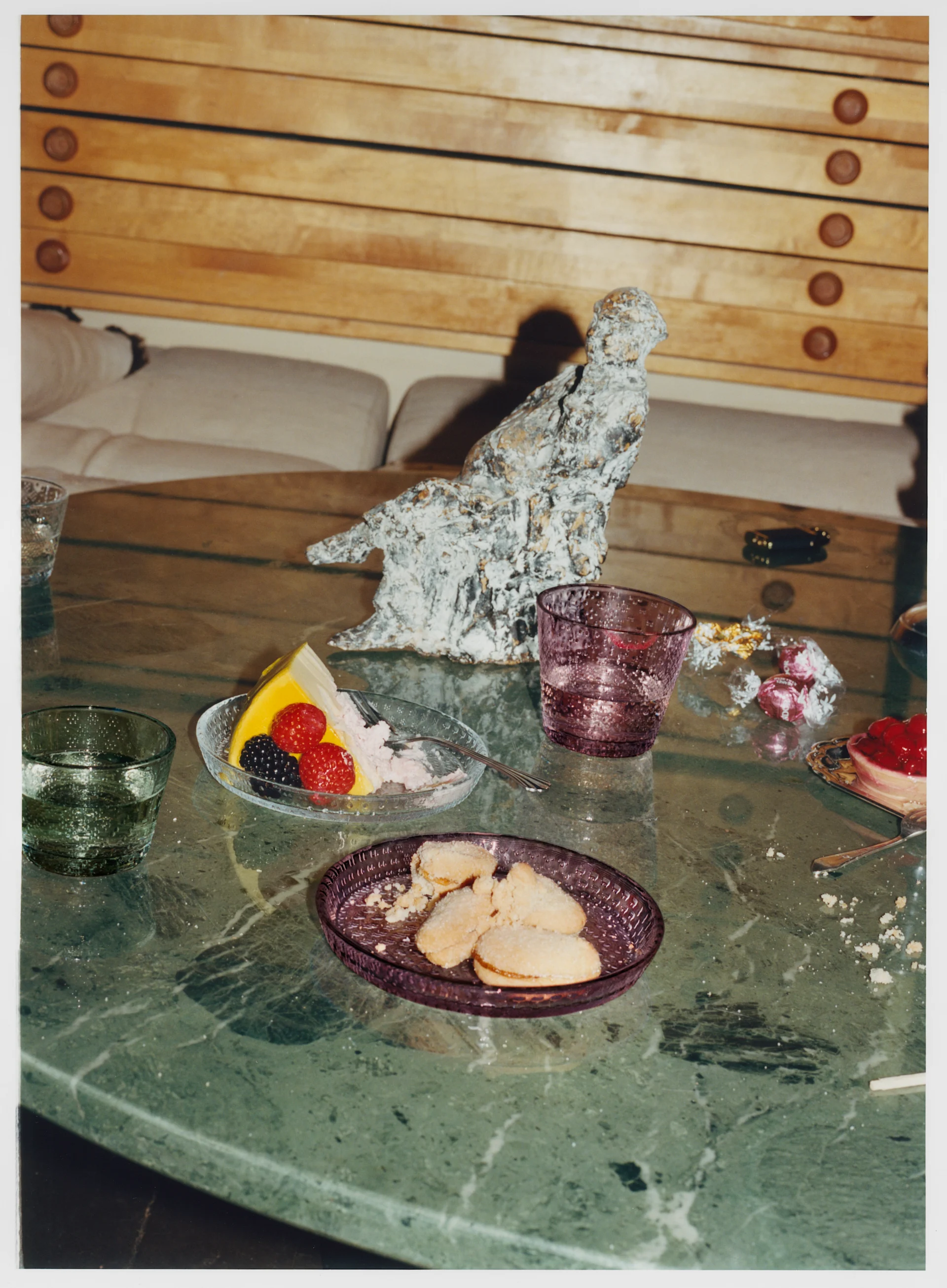 A marble table with a slice of cake, cookies, fruit tart, and glasses. A textured white sculpture is in the background.
