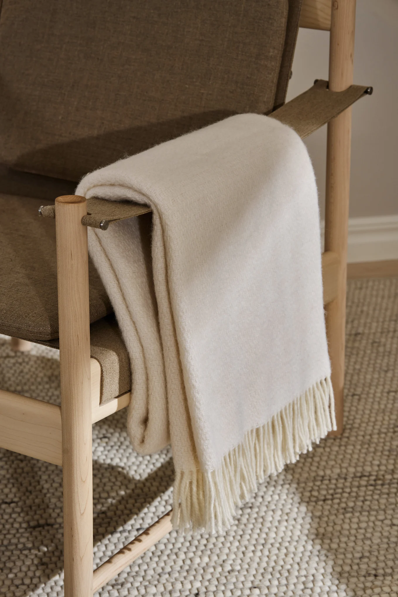 A light off-white fringed blanket draped over a wooden armchair with beige upholstery, on a textured rug.