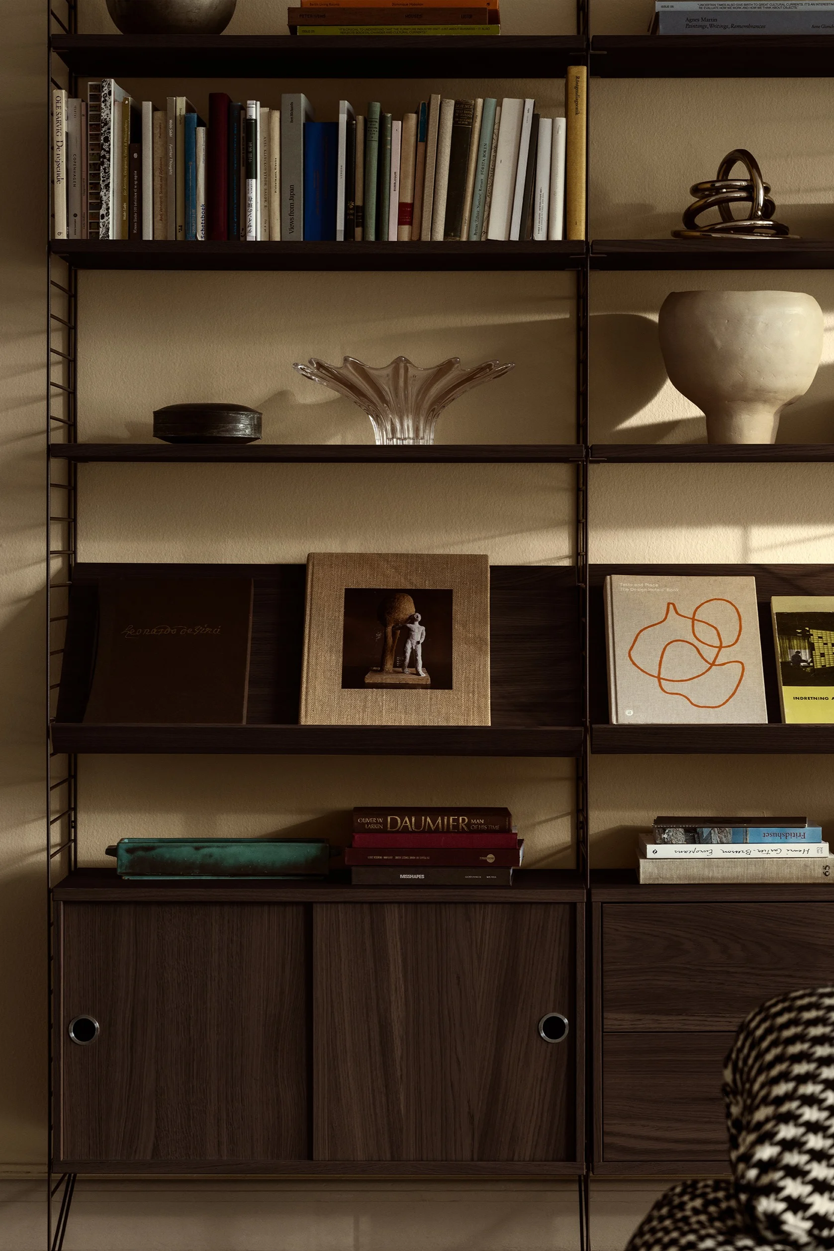 Dark wood modular shelving with books, clear fluted vase, beige ceramic pot, and a metallic knot sculpture.
