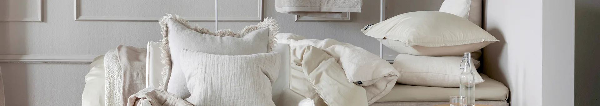 Stacked natural-toned pillows and soft bedding, with a fringed pillow and a glass water bottle.