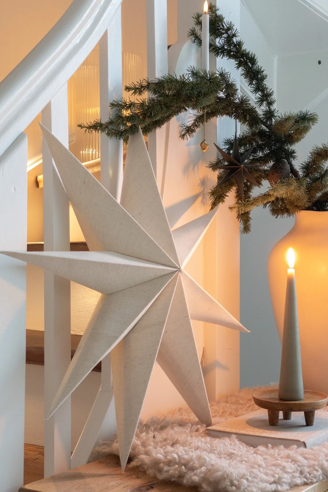 A large fabric Christmas star, evergreen garland with ornaments, and lit candles create a cozy holiday display near a white staircase.