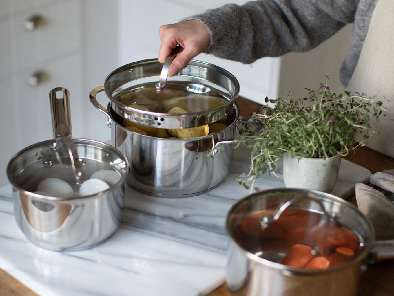 Hand lifting lid from a steaming pot of potatoes, with eggs and carrots in other pots.