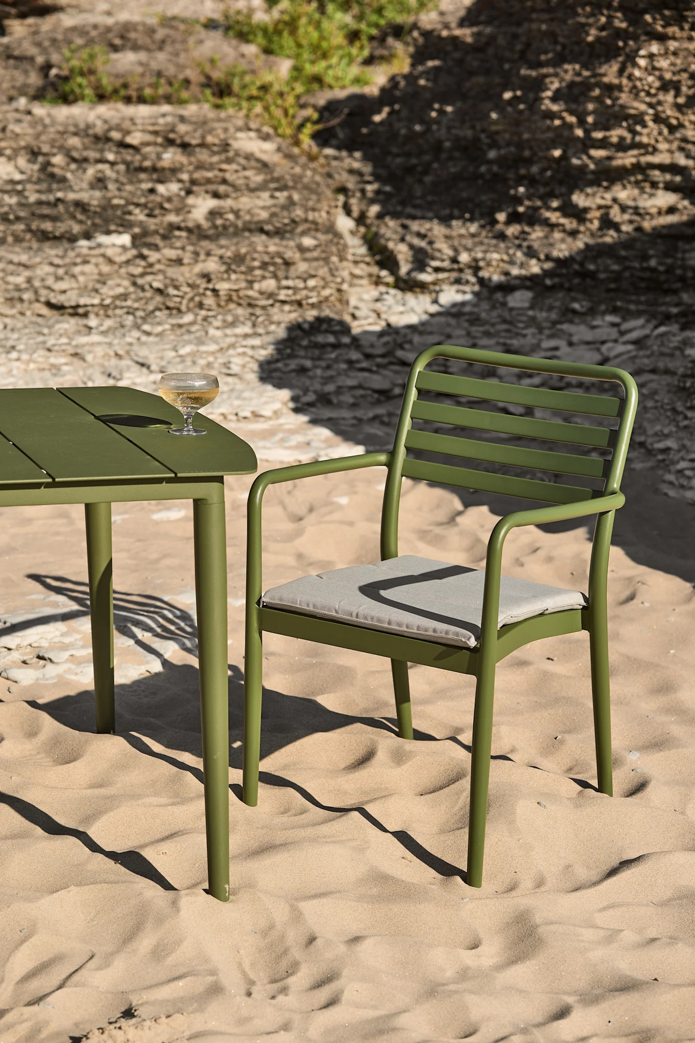 Green metal outdoor table and chair on a sandy beach with a glass of white wine on the table.