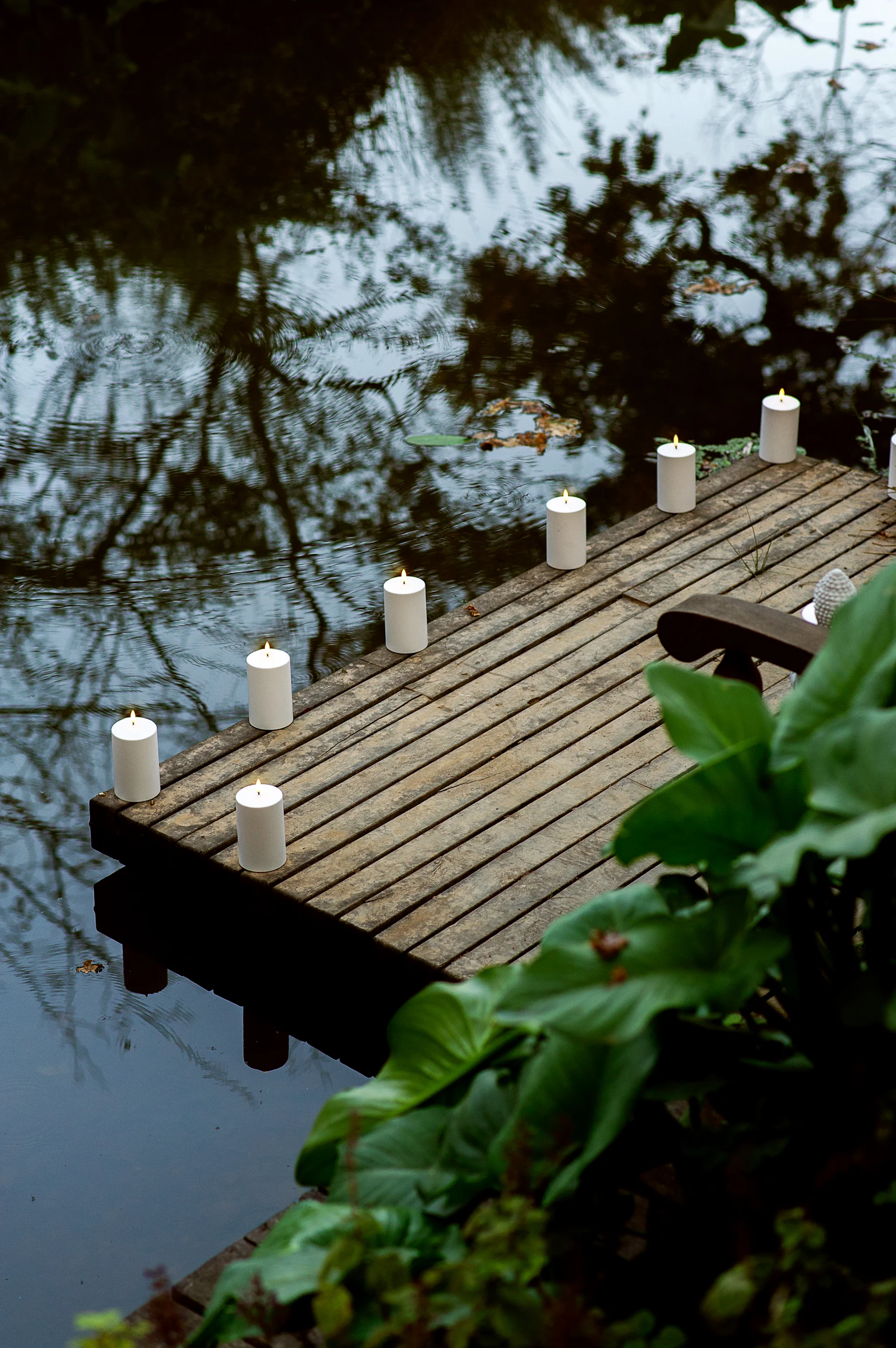 Lit white pillar candles line a wooden dock over a calm pond, with reflections and green foliage.