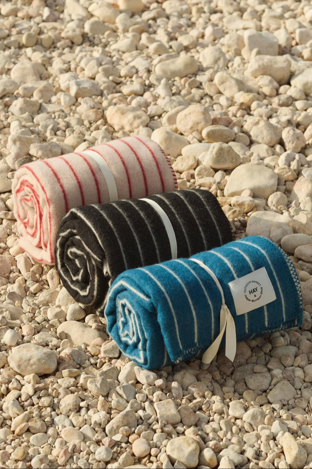 Three rolled-up striped blankets in pink, black, and blue, tied with ribbons on a pebble beach.