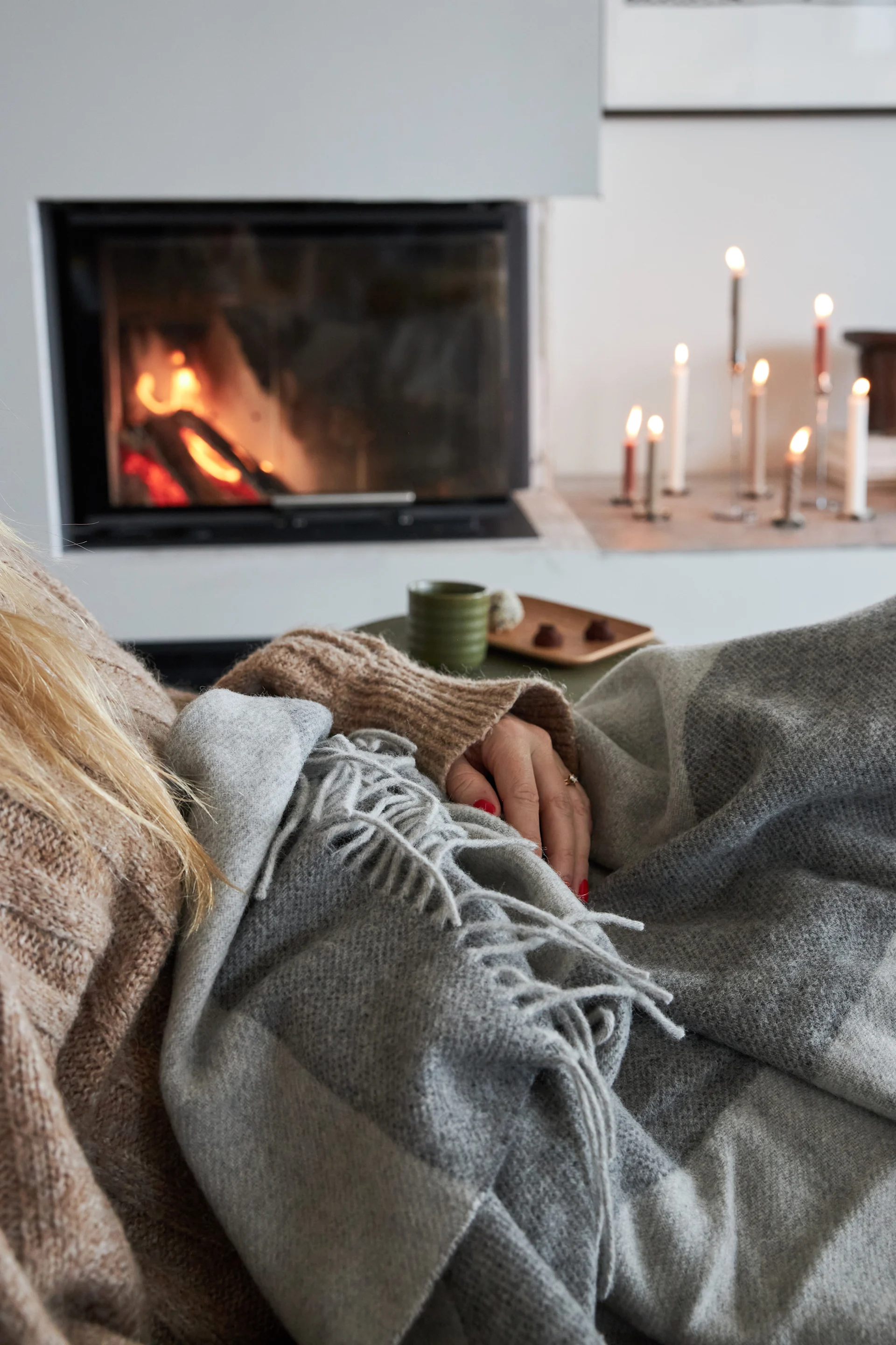 Person cozy under a grey blanket by a fireplace, with lit candles, a cup, and chocolates nearby.