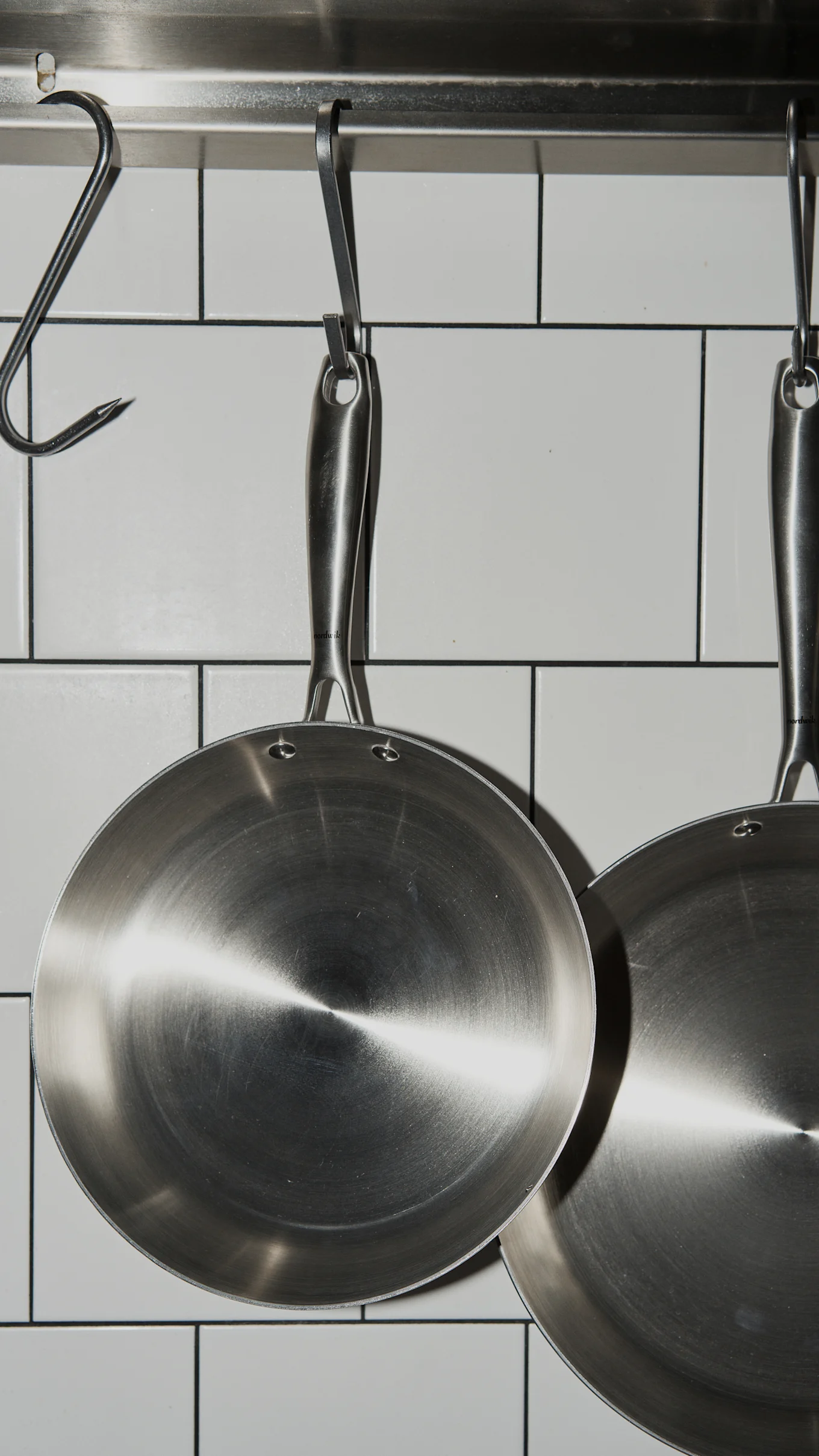 Two shiny stainless steel frying pans hang from hooks on a metal rail against a white tiled wall.