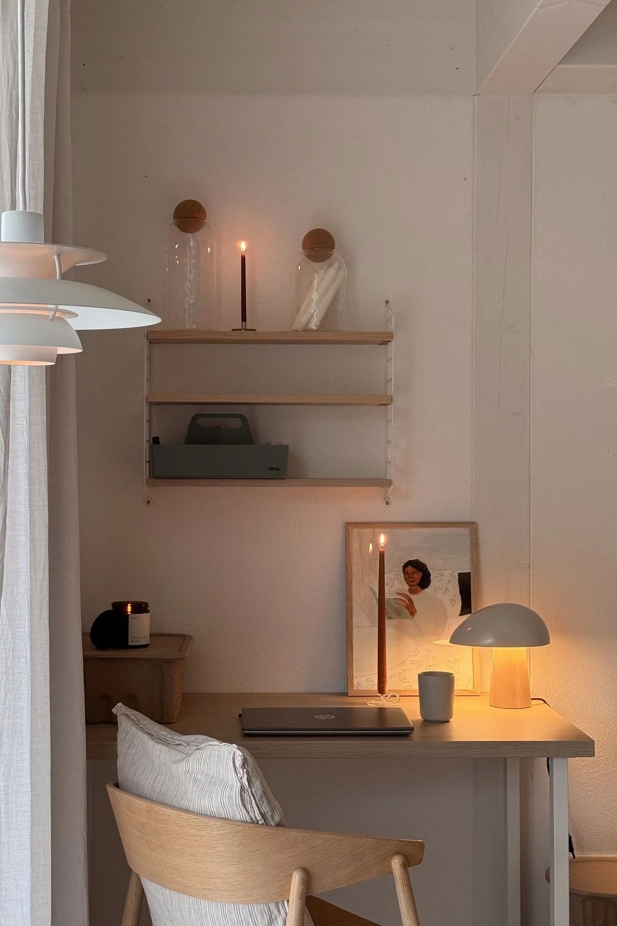 A cozy desk setup with warm lighting, a closed laptop, two lit candles, a modern lamp, and a framed artwork.