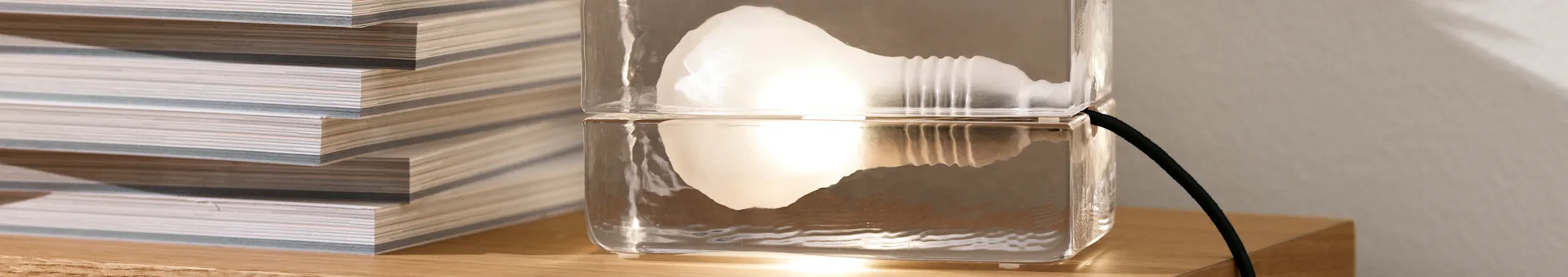 A unique clear lamp, made of two stacked rectangular blocks, with a glowing internal light, next to a stack of magazines on a wooden surface.