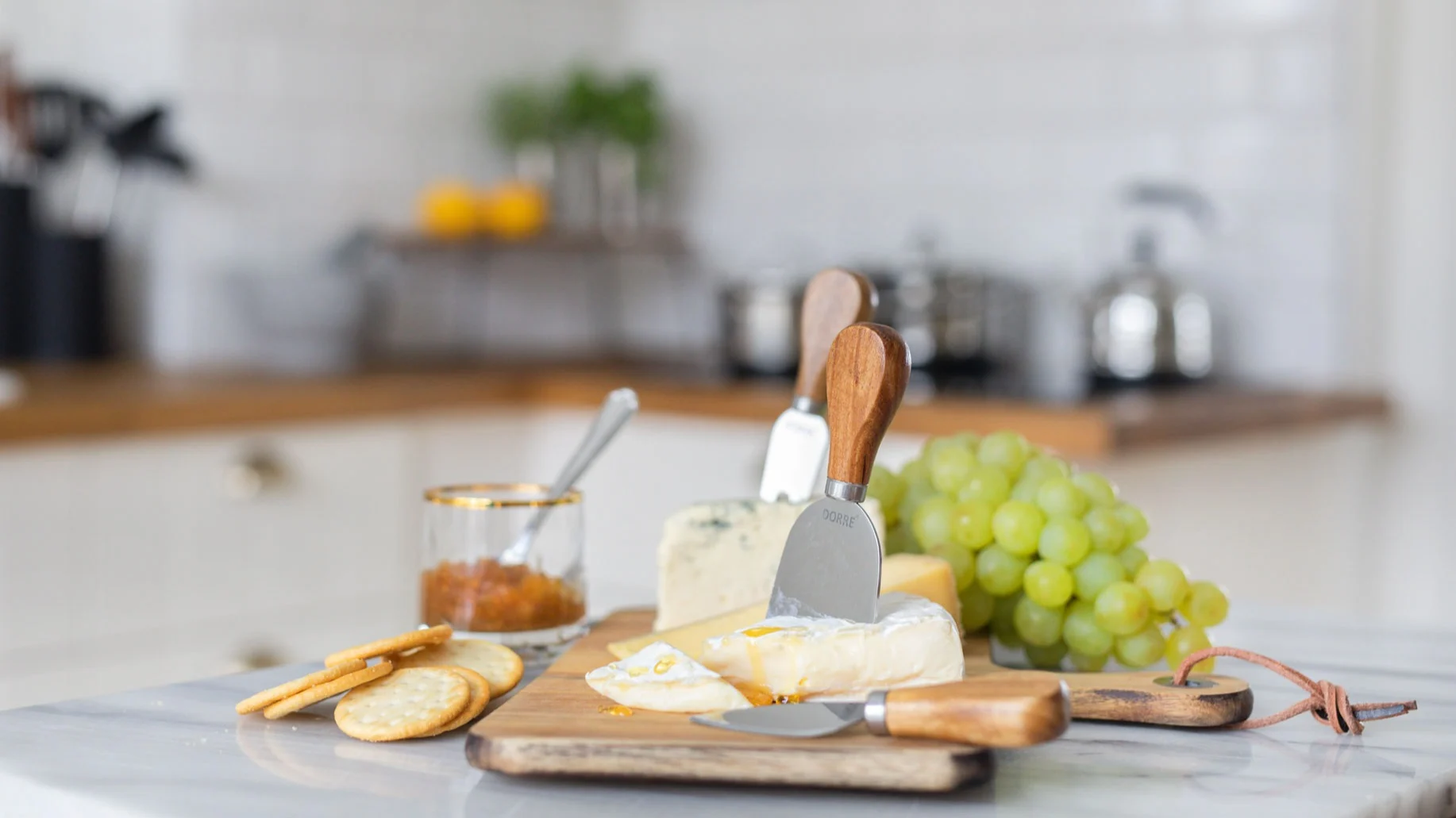 A wooden board with sliced Brie, blue cheese, crackers, green grapes, and jam, set on a marble kitchen counter.