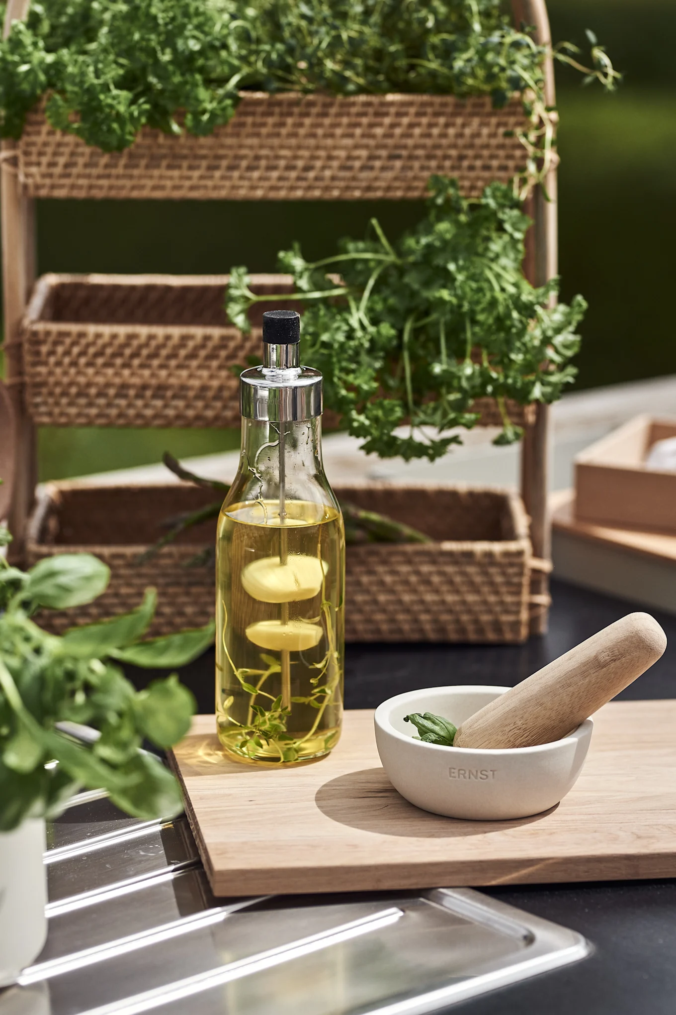 Oil infuser bottle with herbs, white mortar with basil and wooden pestle on a board. Woven shelves with herbs in background.