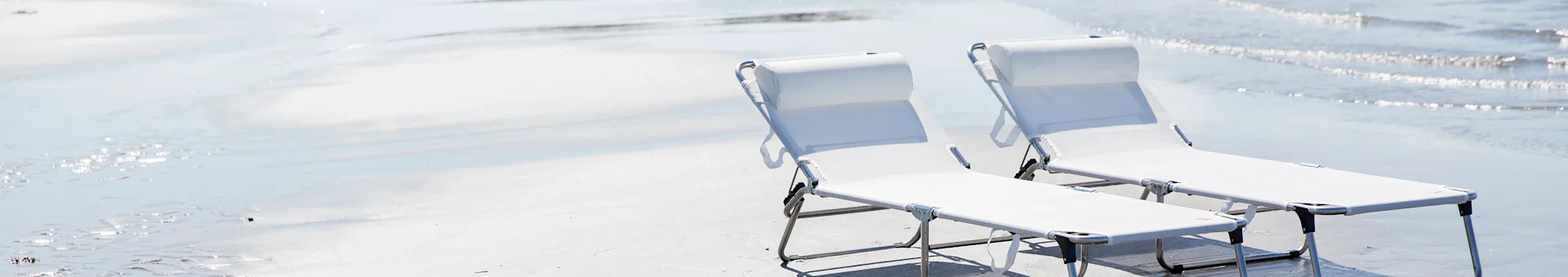 Two white portable lounge chairs on a bright beach with shimmering water.