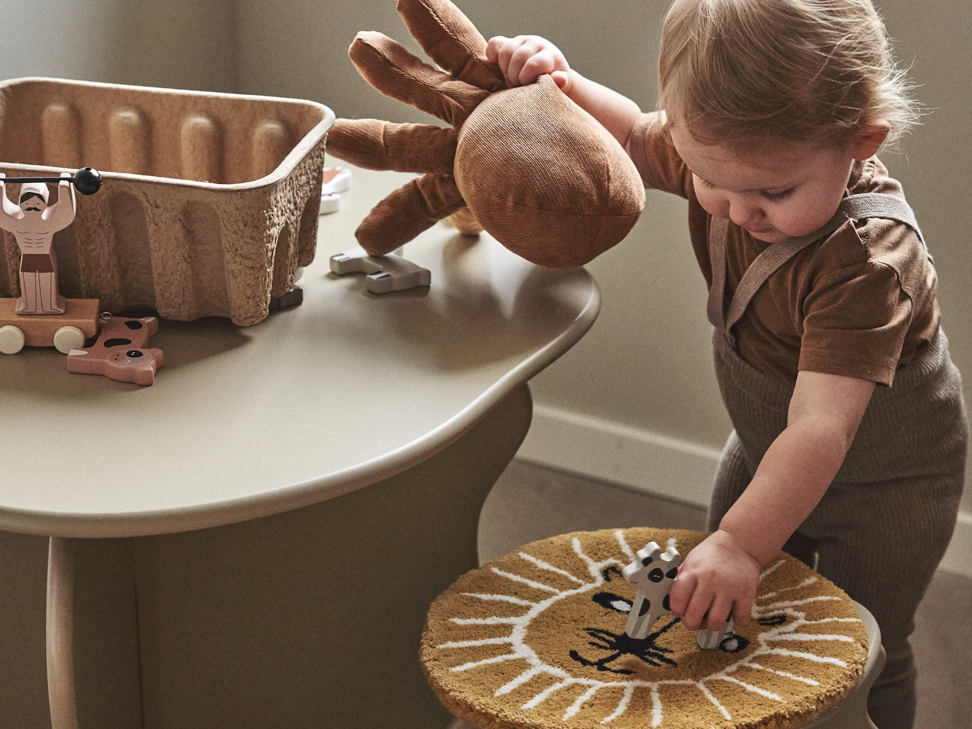 Toddler plays with a brown stuffed animal, placing it on a lion-patterned stool with other wooden toys on a table.
