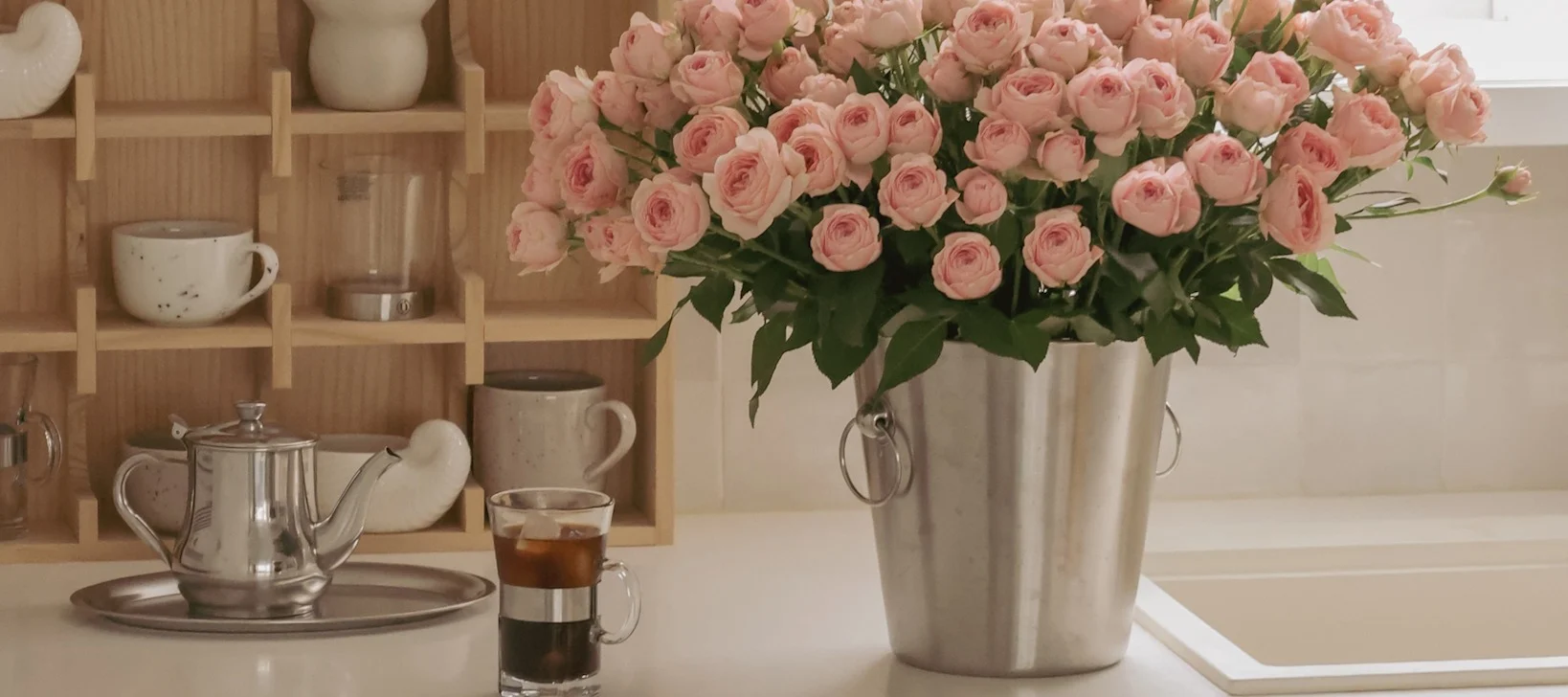 A large bouquet of pink roses in a silver bucket sits on a kitchen counter with a teapot and iced coffee.