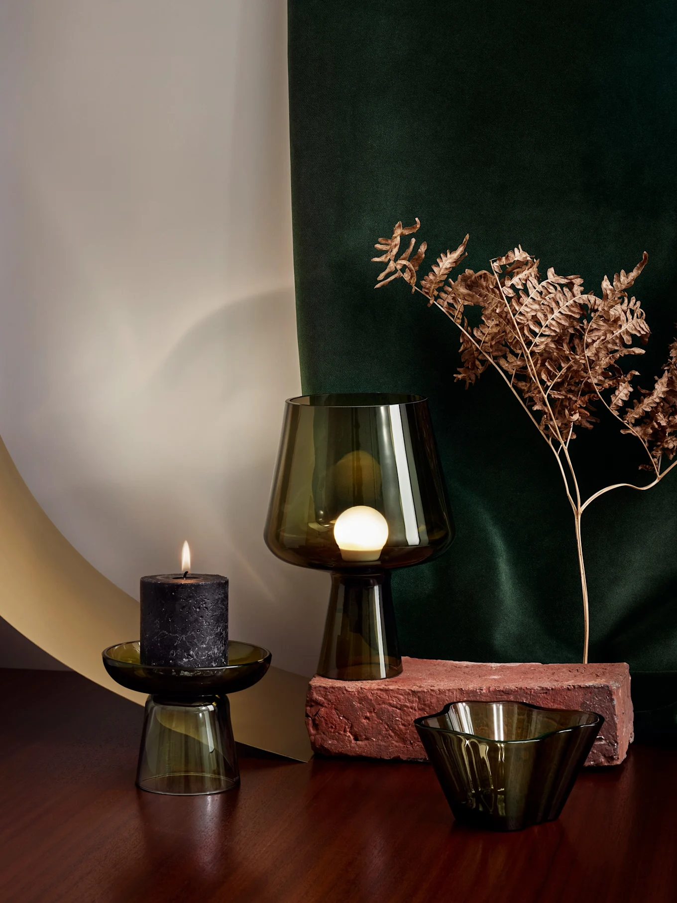 A dark green glass lamp on a brick, a lit black candle on a glass holder, and a glass bowl on a wooden surface, with a dried fern and green drape in the background.