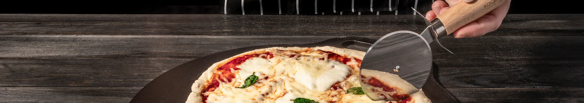 Close-up of a person cutting a pizza with cheese, tomato sauce, and basil on a dark wooden table.