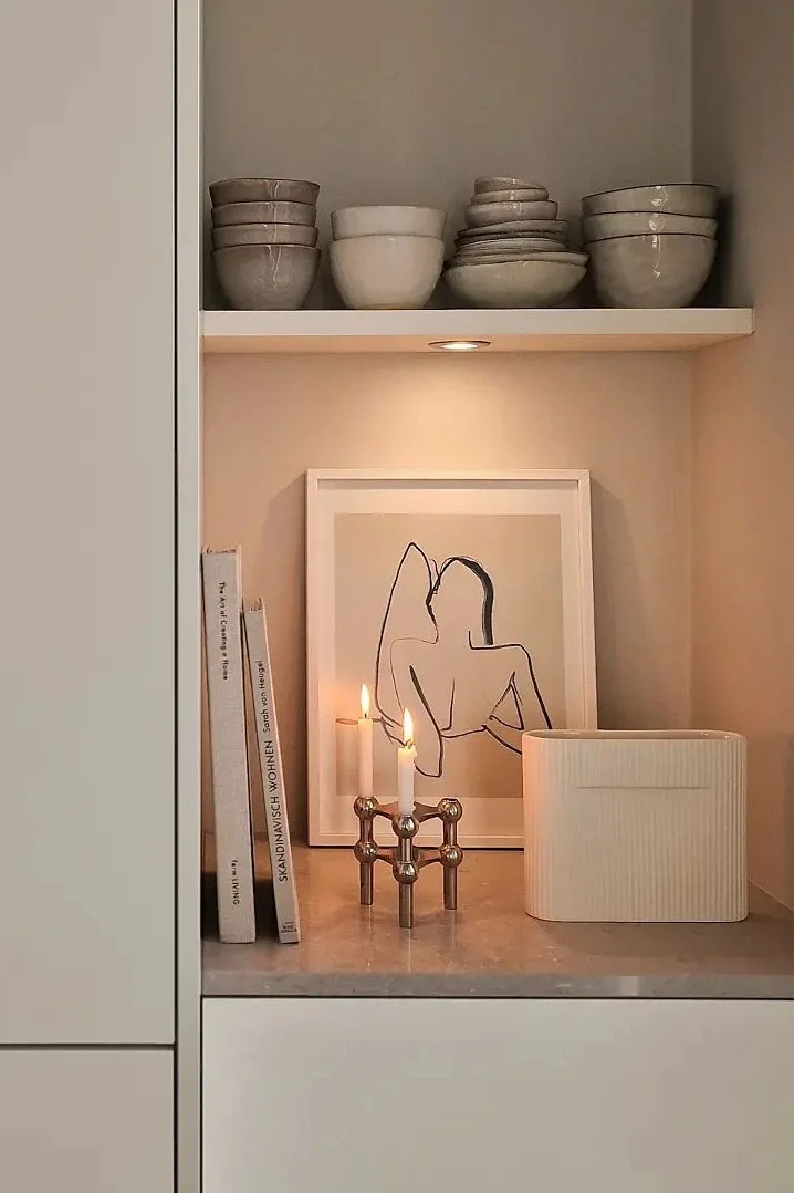 A cozy shelf with ceramic bowls, books, framed art, lit candles in a brass holder, and a ridged vase.