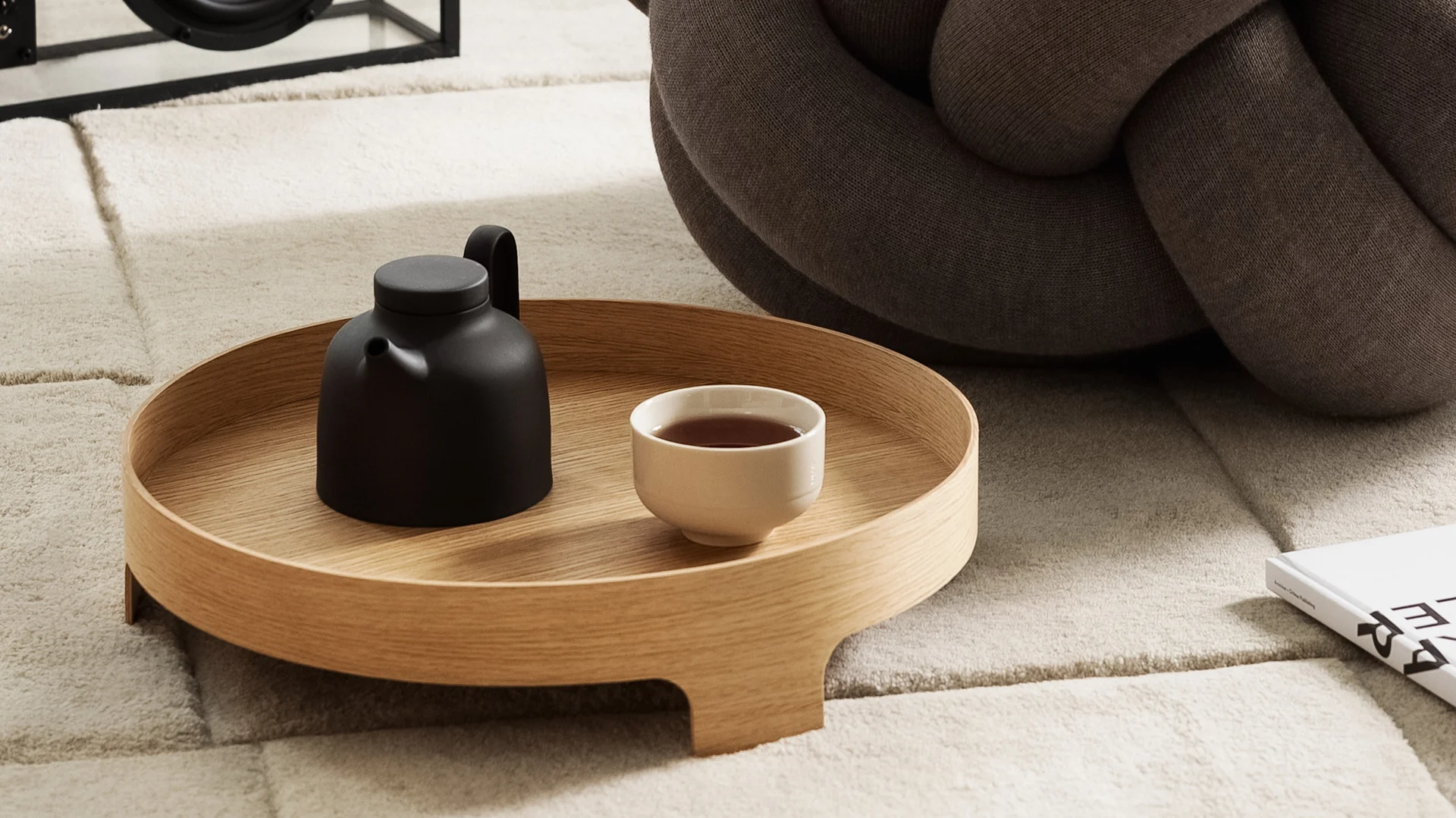 A black teapot and a cup of tea on a round wooden tray, resting on a beige textured rug next to a brown knotted cushion.