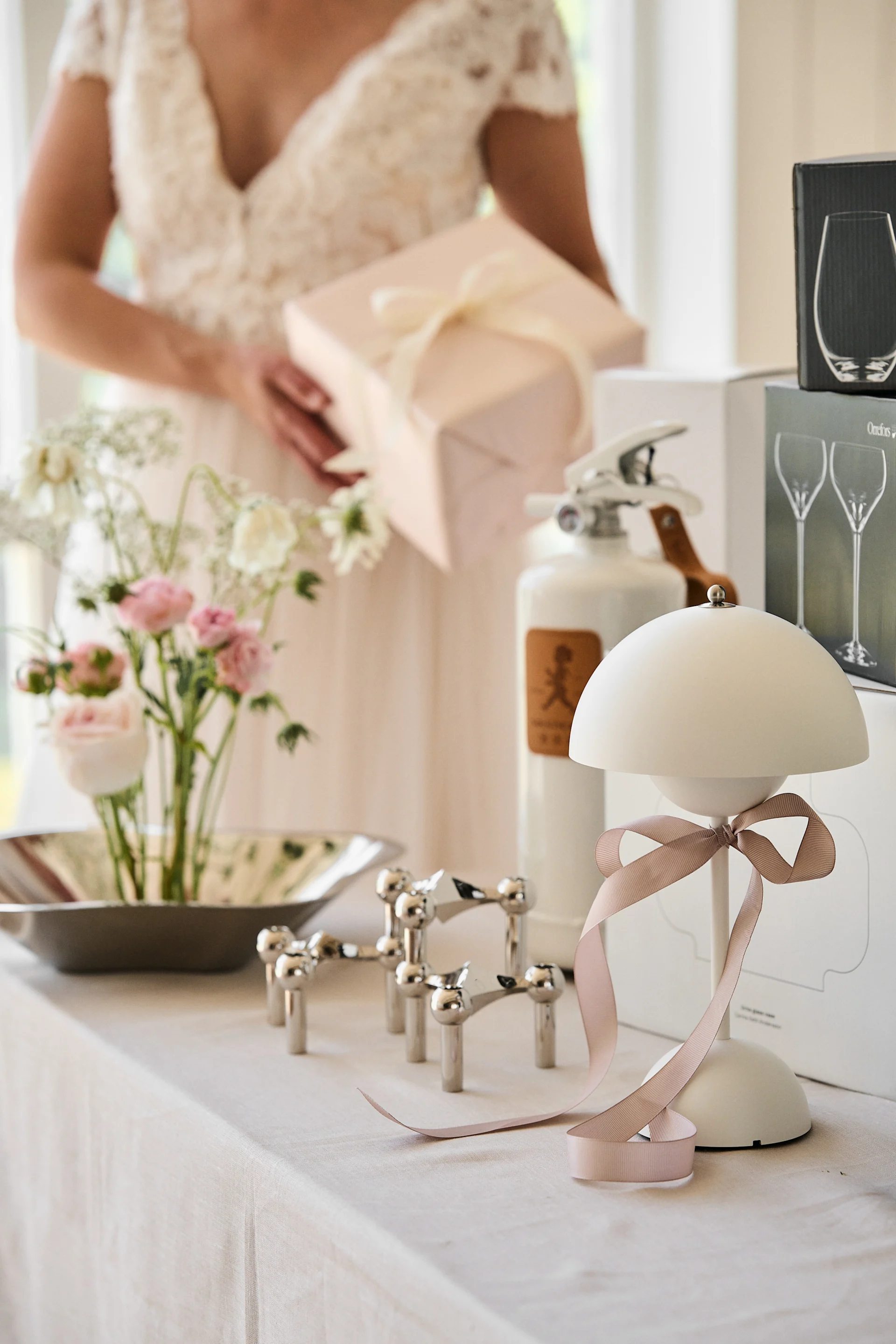 Person in light dress holds pink gift box behind table with flowers, chrome candelabra, white lamp with pink ribbon, and gift boxes.