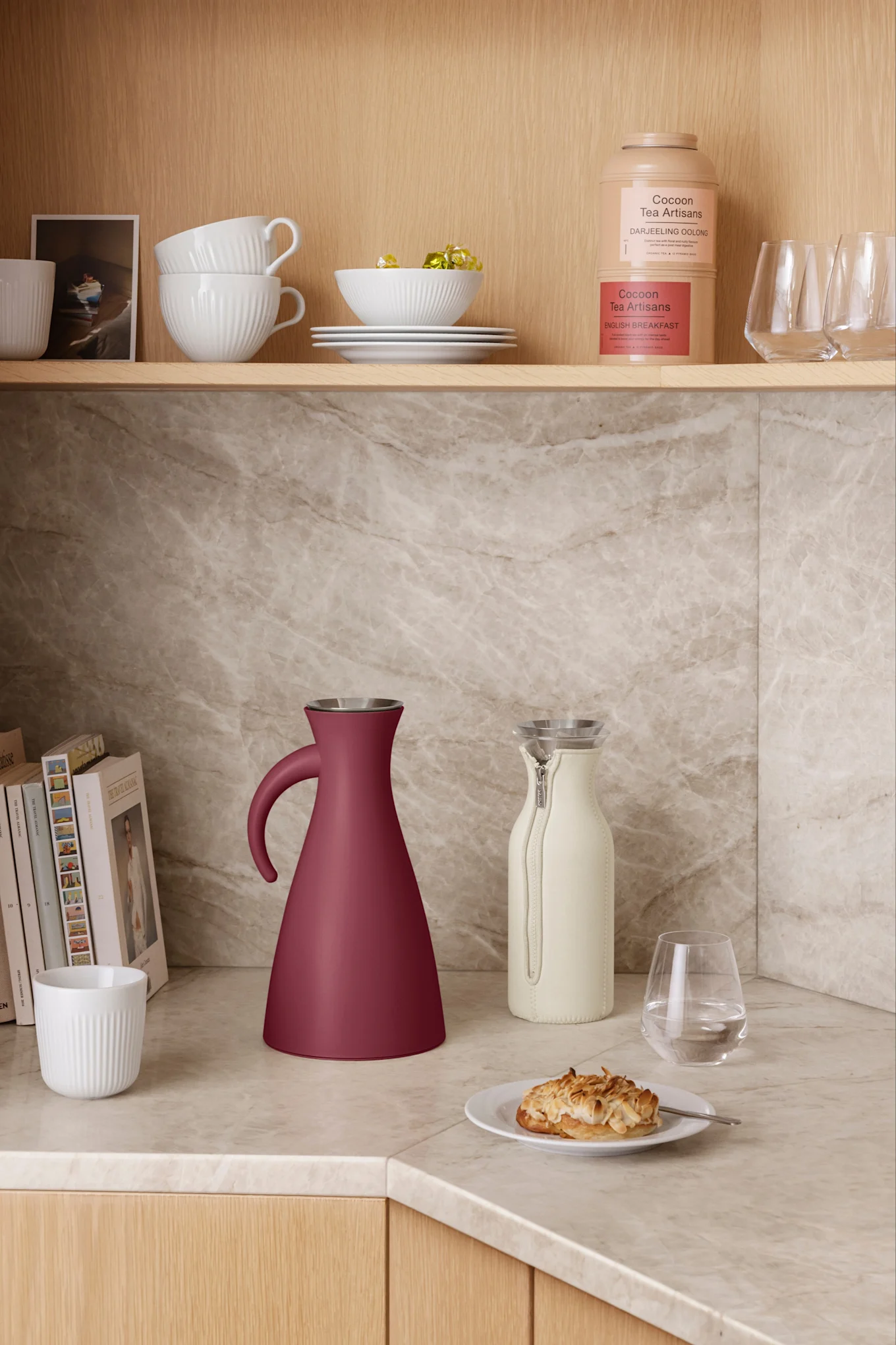 A dark red conical thermos and a light cream pitcher on a marble counter. White bowls, mugs, tea containers are on shelves above.
