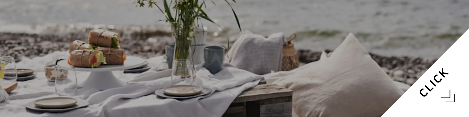A serene beach picnic with wrapped food, tableware, a vase of flowers, and cushions on a pebble shore by the sea.