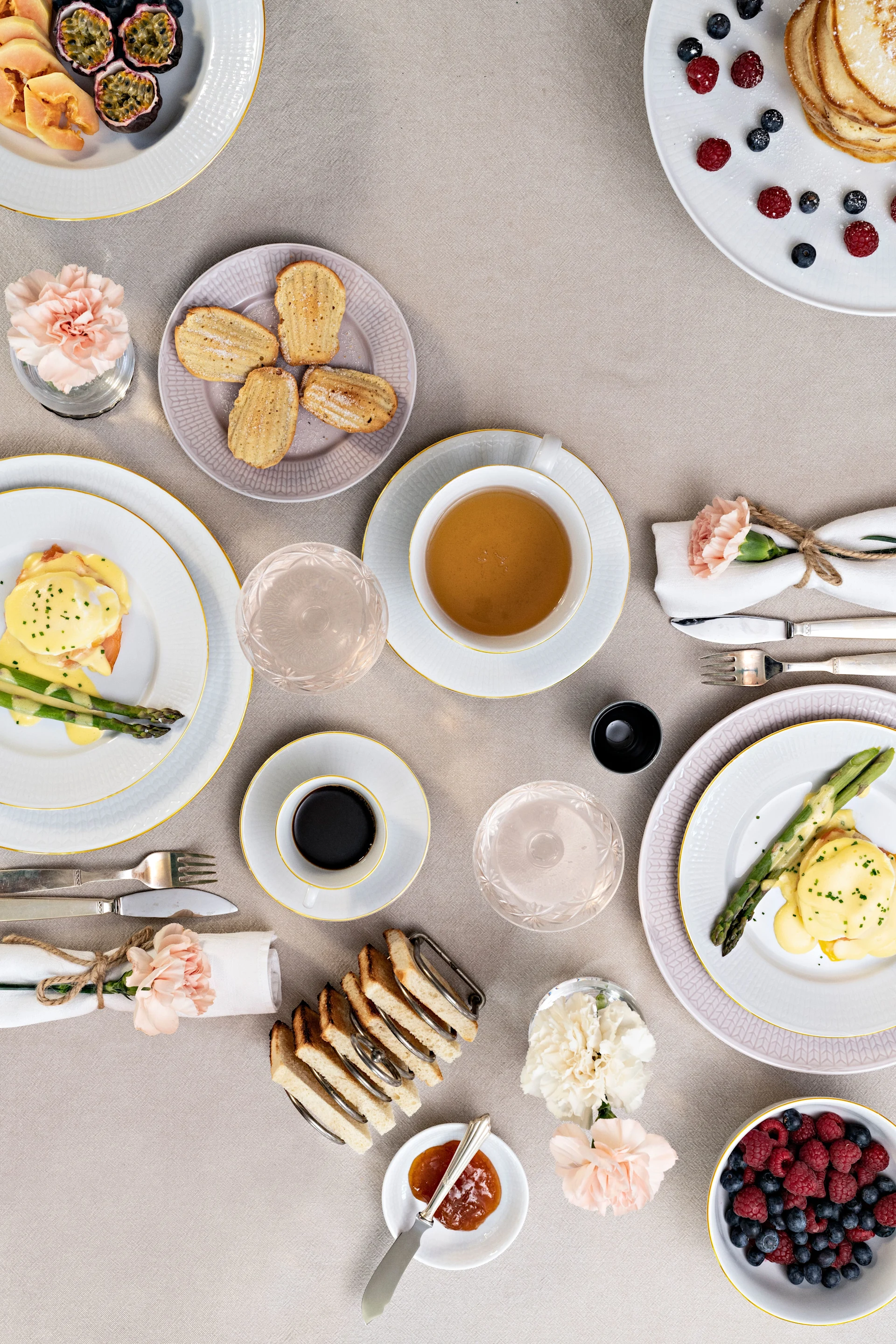 Brunch spread on beige linen: Eggs Benedict, pancakes, fresh fruit, madeleines, toast, coffee, and tea.