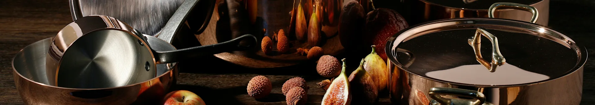 Shiny copper pots and pans with lychees, figs, and an apple on a dark wooden surface.