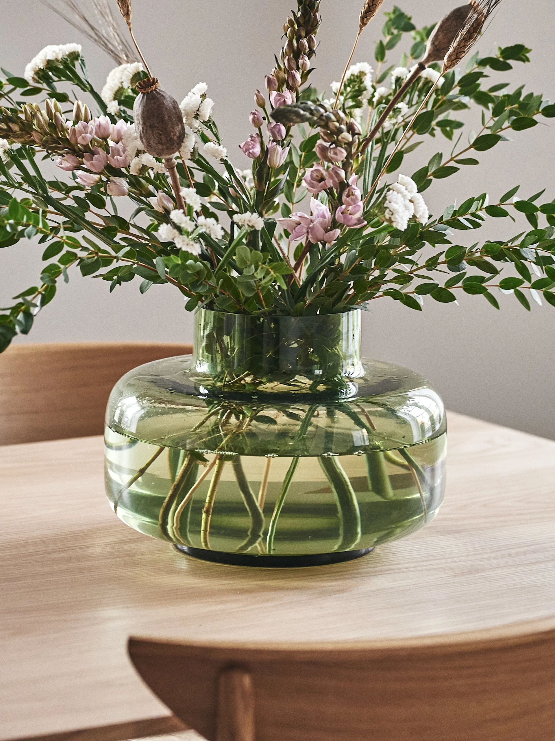 Mixed bouquet of pink and white flowers, green leaves, and dried elements in a squat green glass vase on a wooden table.