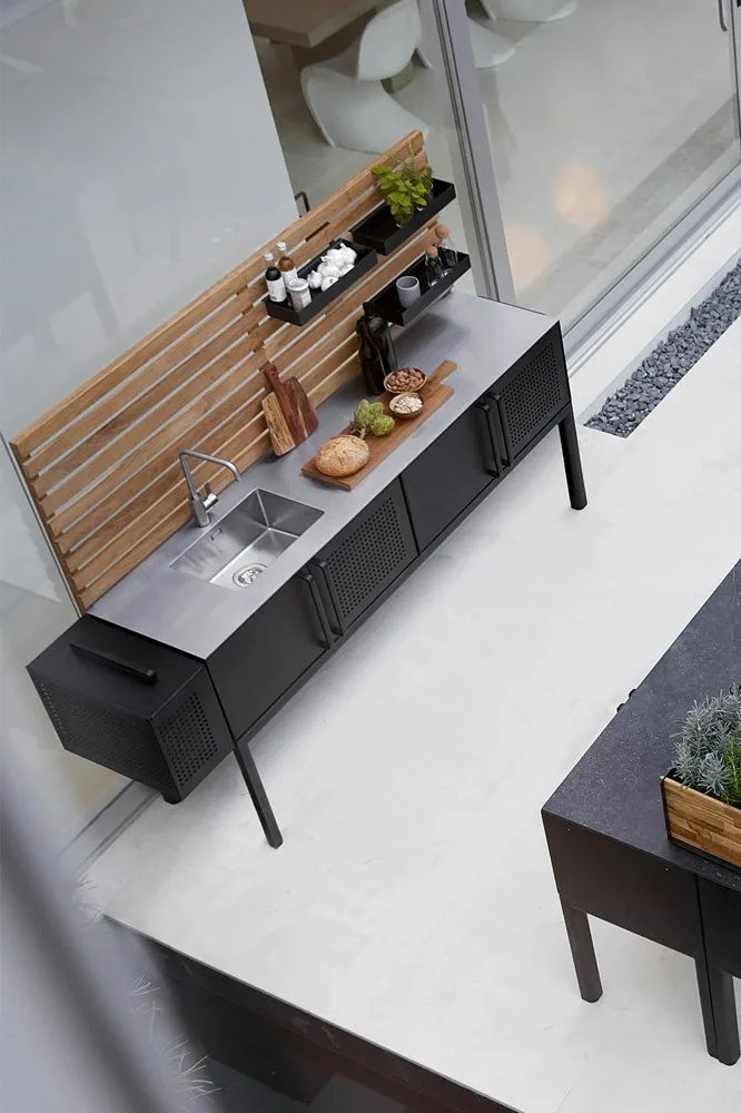 Modern outdoor kitchen with stainless steel sink, black cabinets, wooden back panel, and shelves holding spices and food.