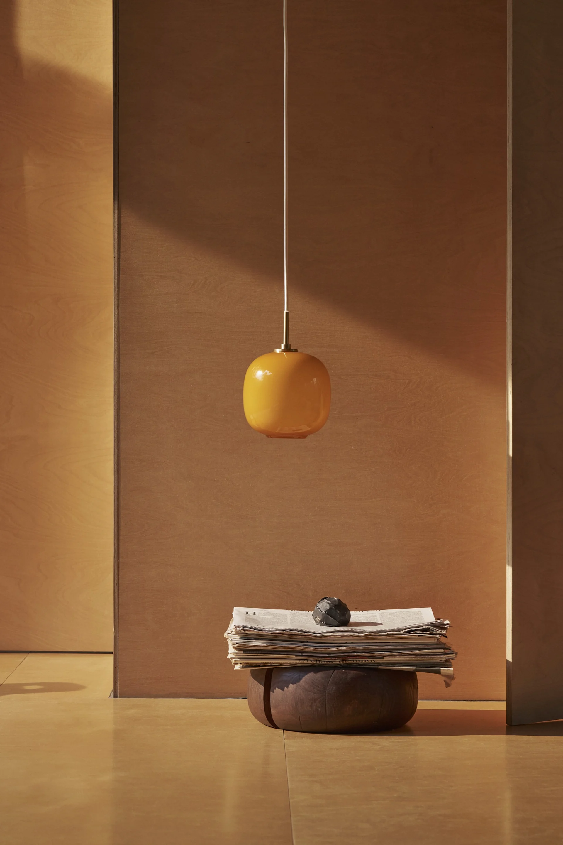 An orange oval pendant light hangs above a stack of newspapers and a dark decorative object on a wooden base in a minimalist room.