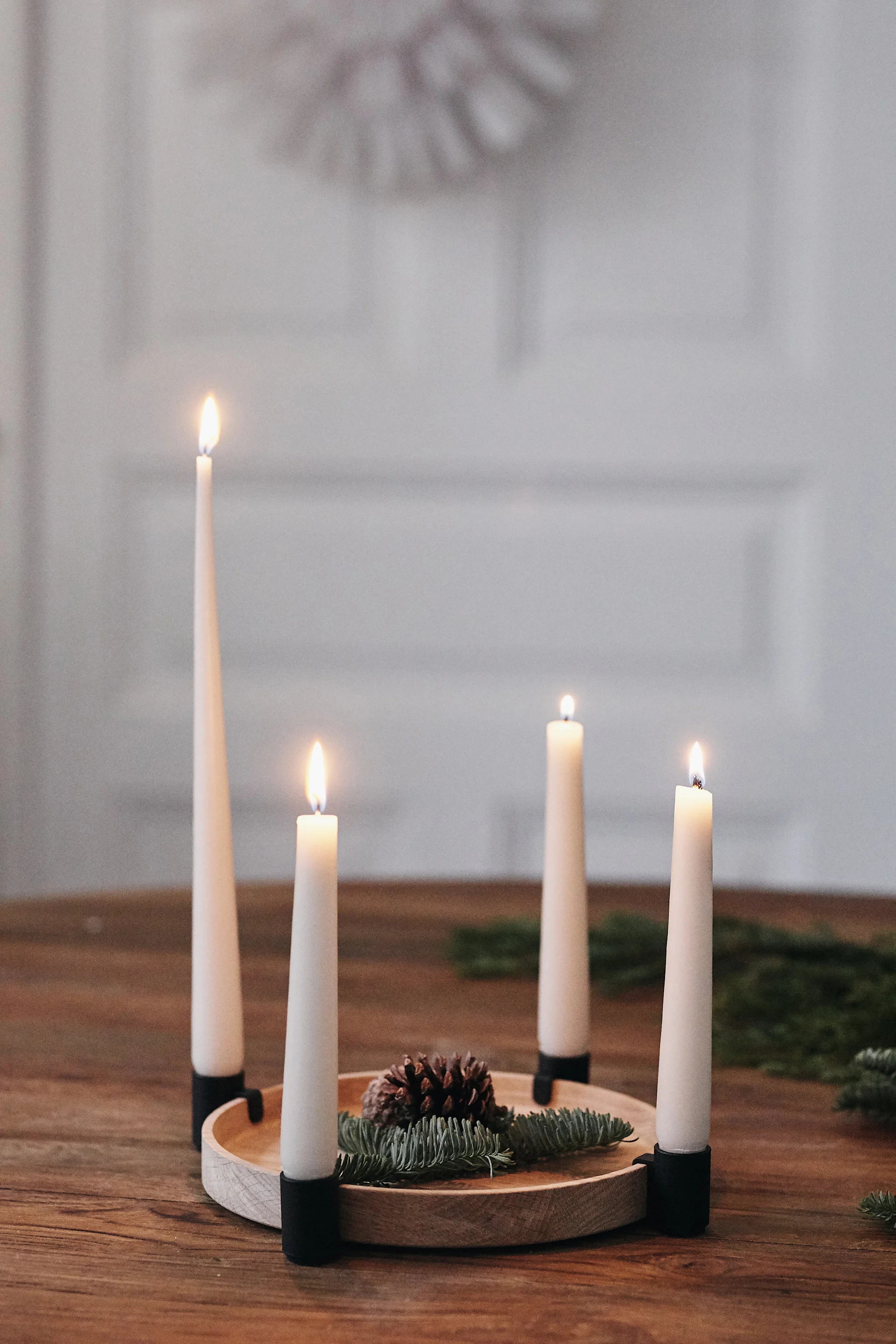 Four white lit candles, arranged on a wooden tray with a pinecone and evergreen branches, on a wooden table.