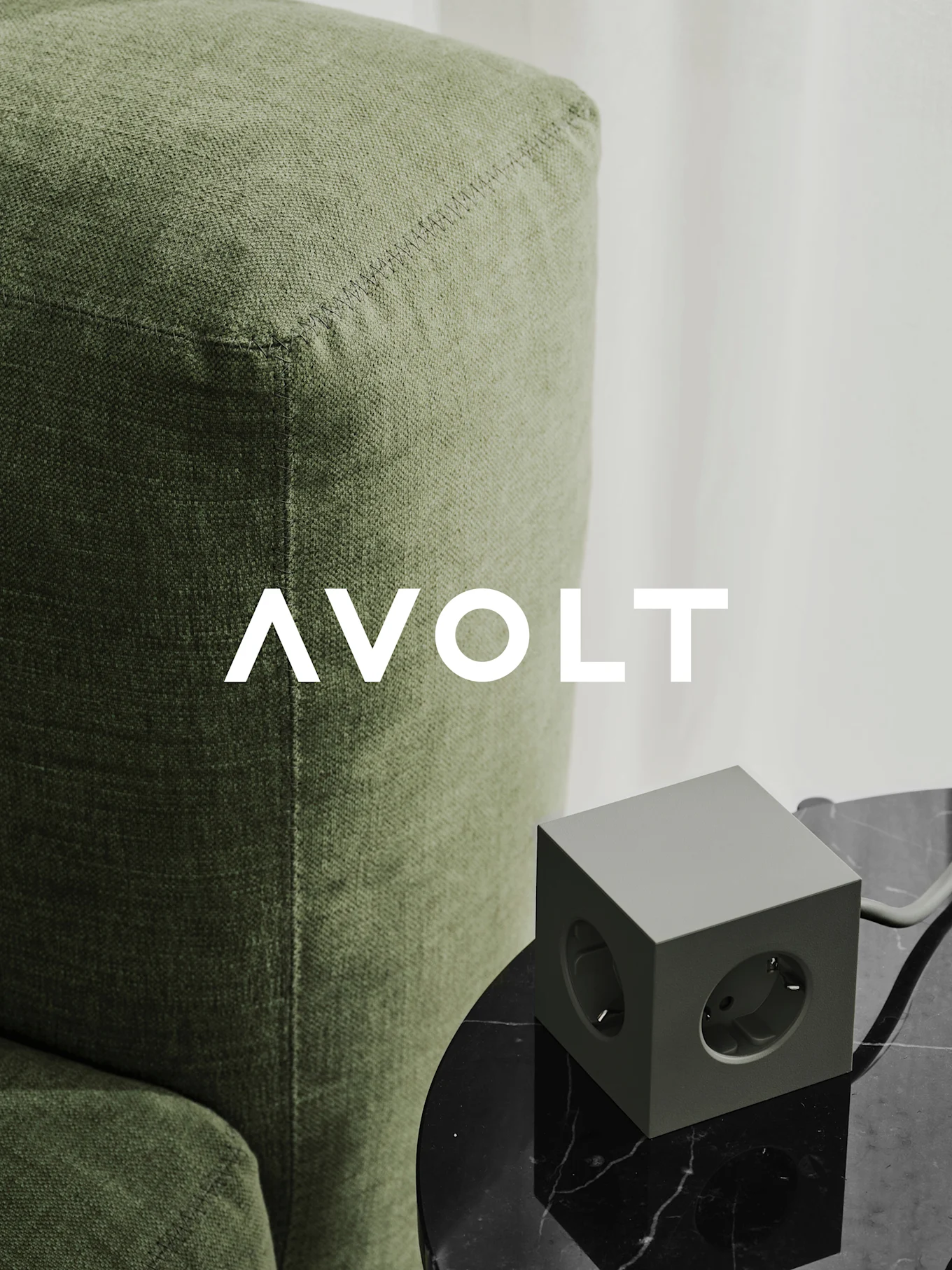 A green cube power strip with three outlets on a black marble table next to a green sofa. AVOLT logo.