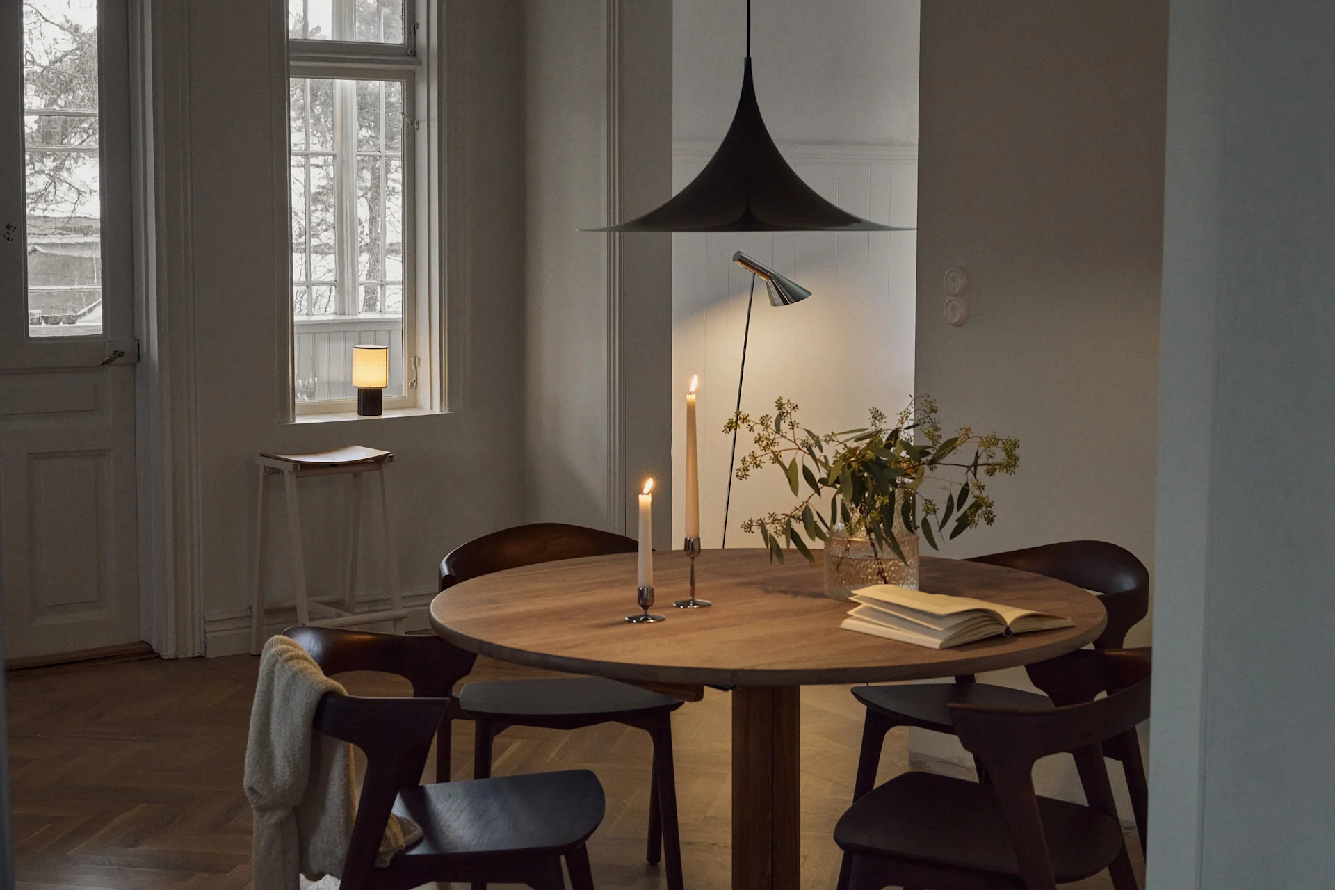 A cozy dining room with a round wooden table, dark chairs, lit candles, greenery, and a bell-shaped pendant lamp.