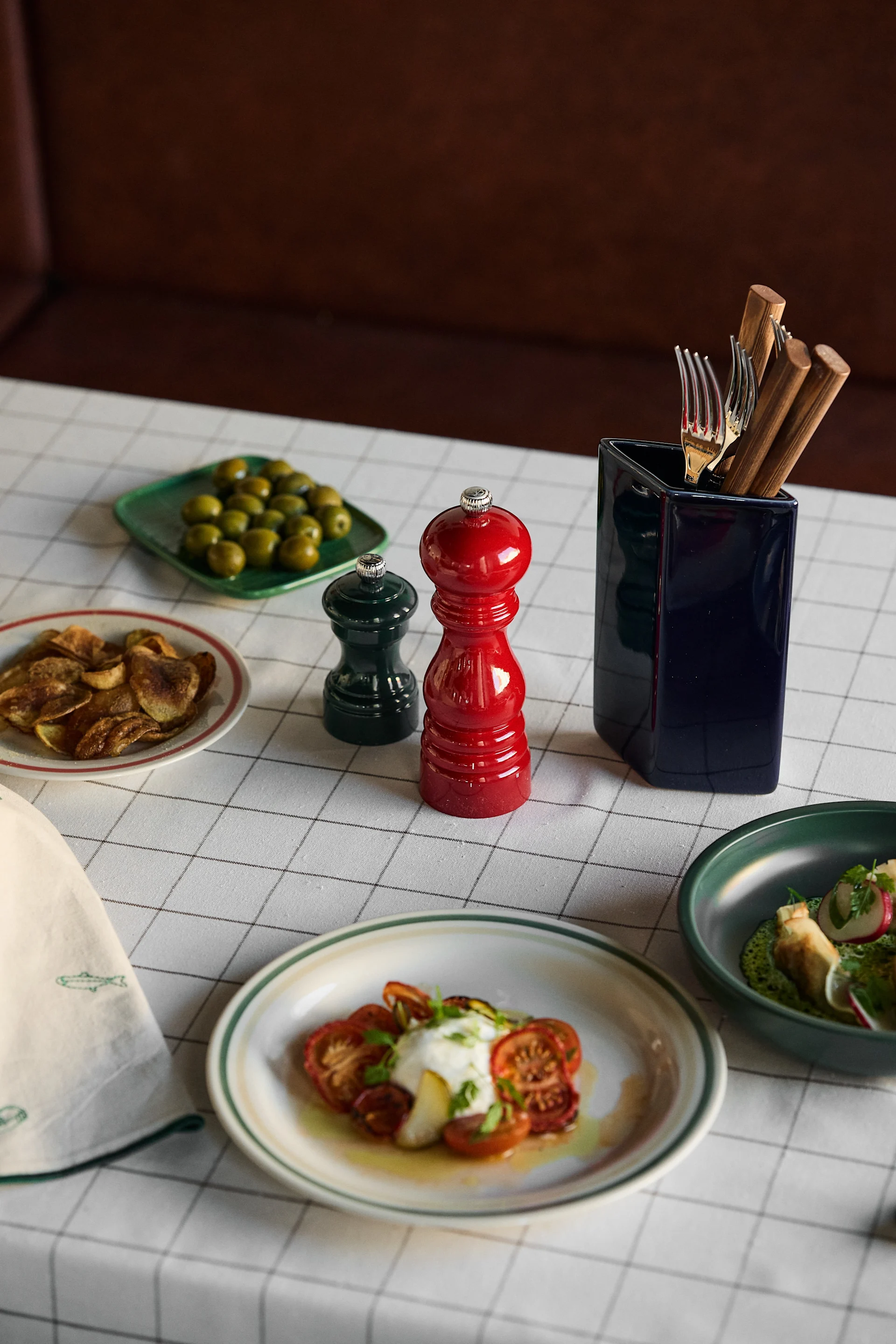 A table set with various dishes including burrata and tomatoes, olives, chips, and utensils, featuring red and green pepper grinders.