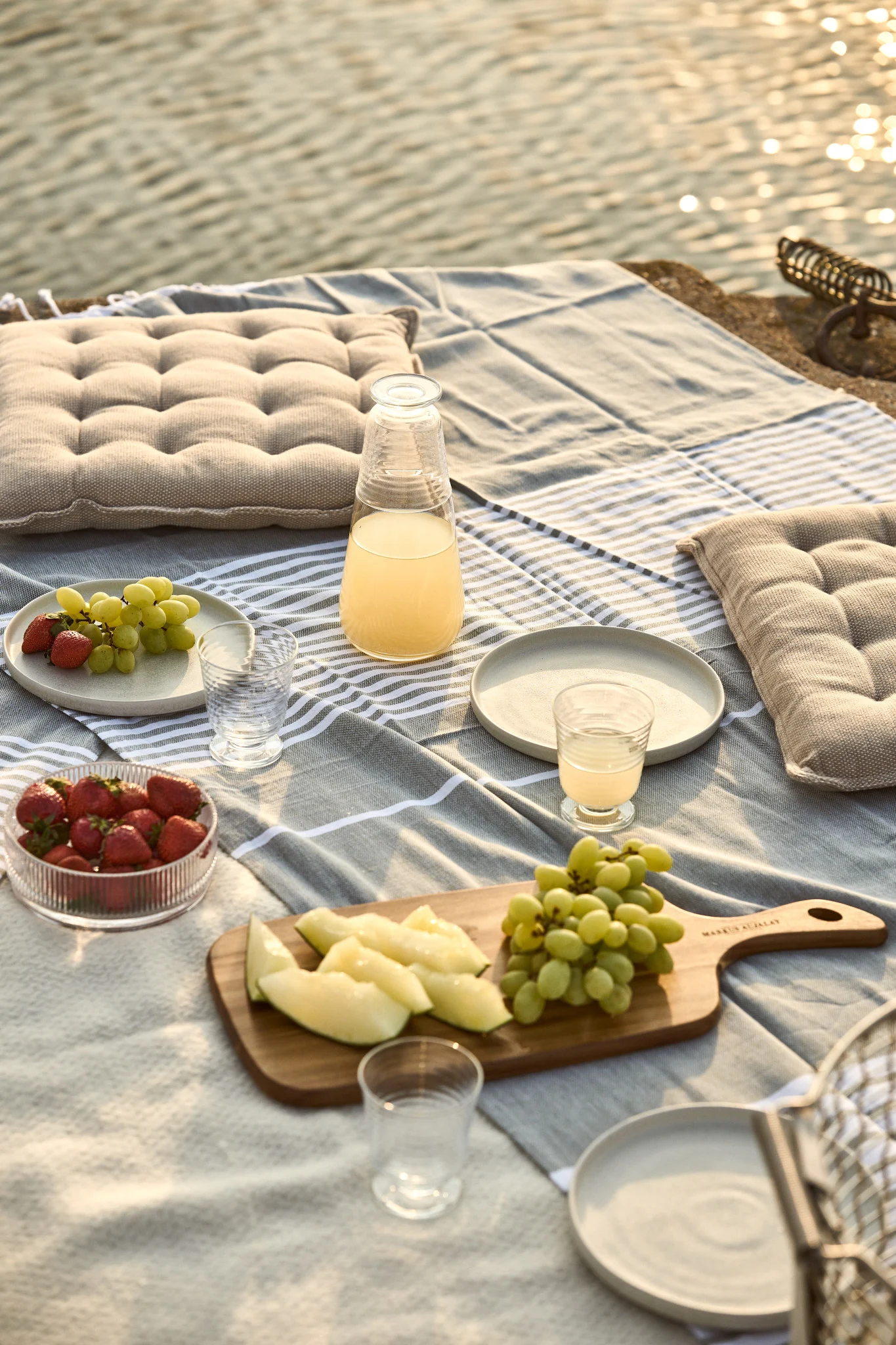 Picnic by the water with a striped blanket, cushions, juice, strawberries, grapes, and melon slices.