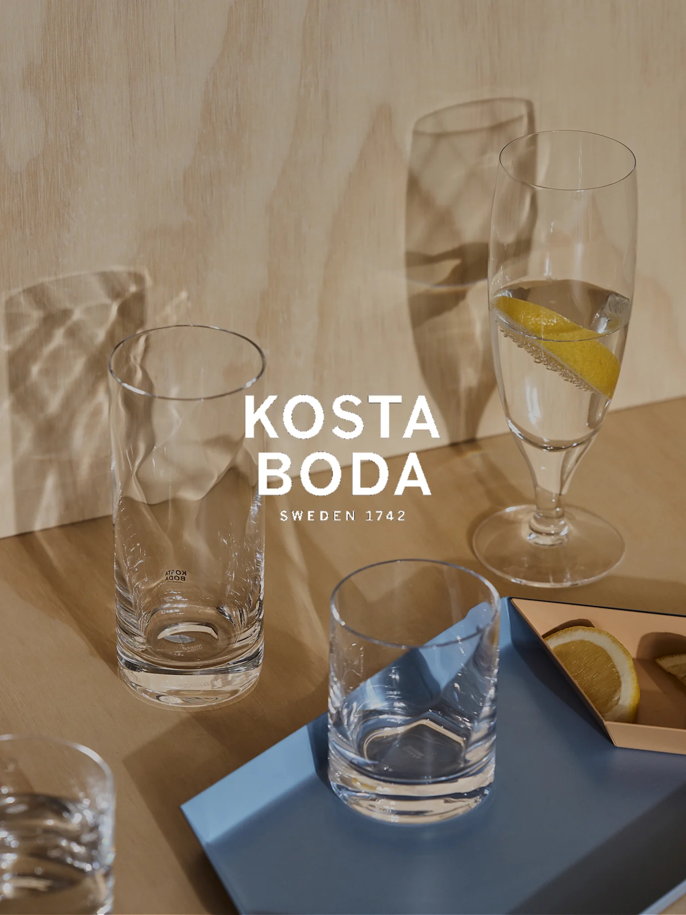 Various clear glass drinking glasses, some with water and lemon, on a light wood surface with blue and orange trays.