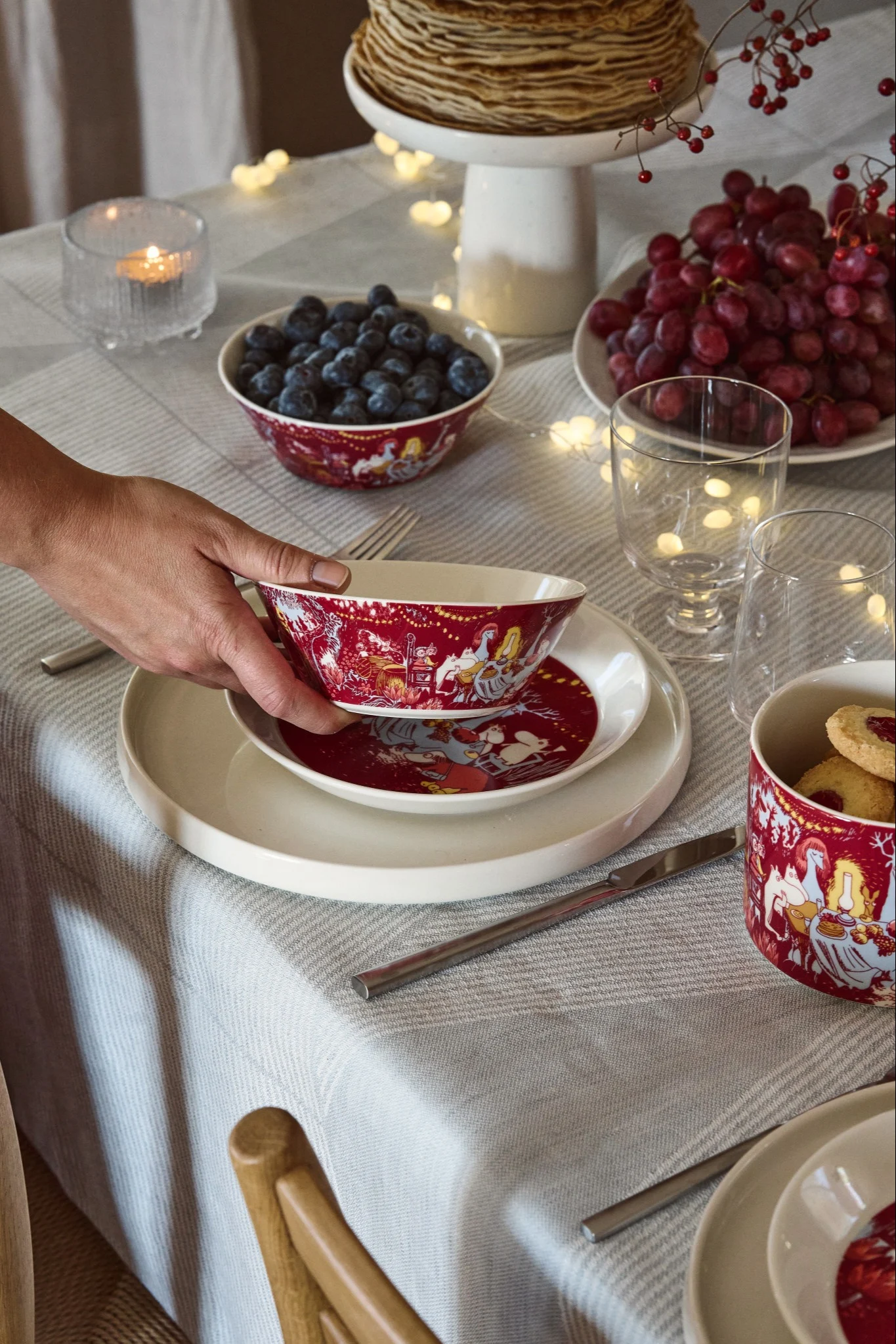 Hand placing a red festive bowl on a white plate at a table set with pancakes, blueberries, grapes, candles, and fairy lights.