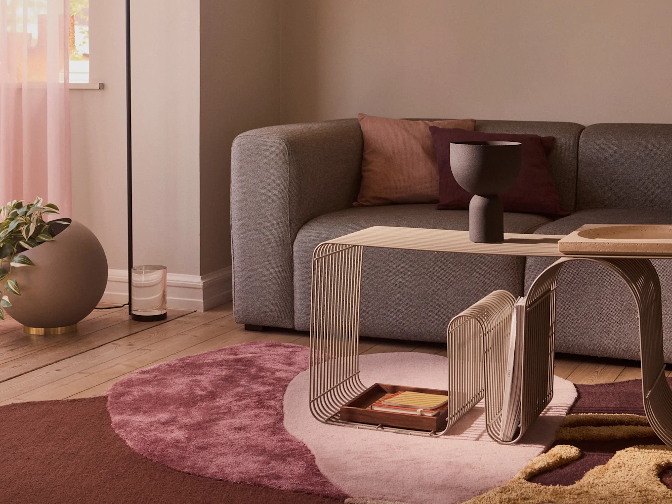 A modern living room with a grey sofa, two minimalist metal side tables, a circular multi-toned rug, and a large plant in a grey pot.