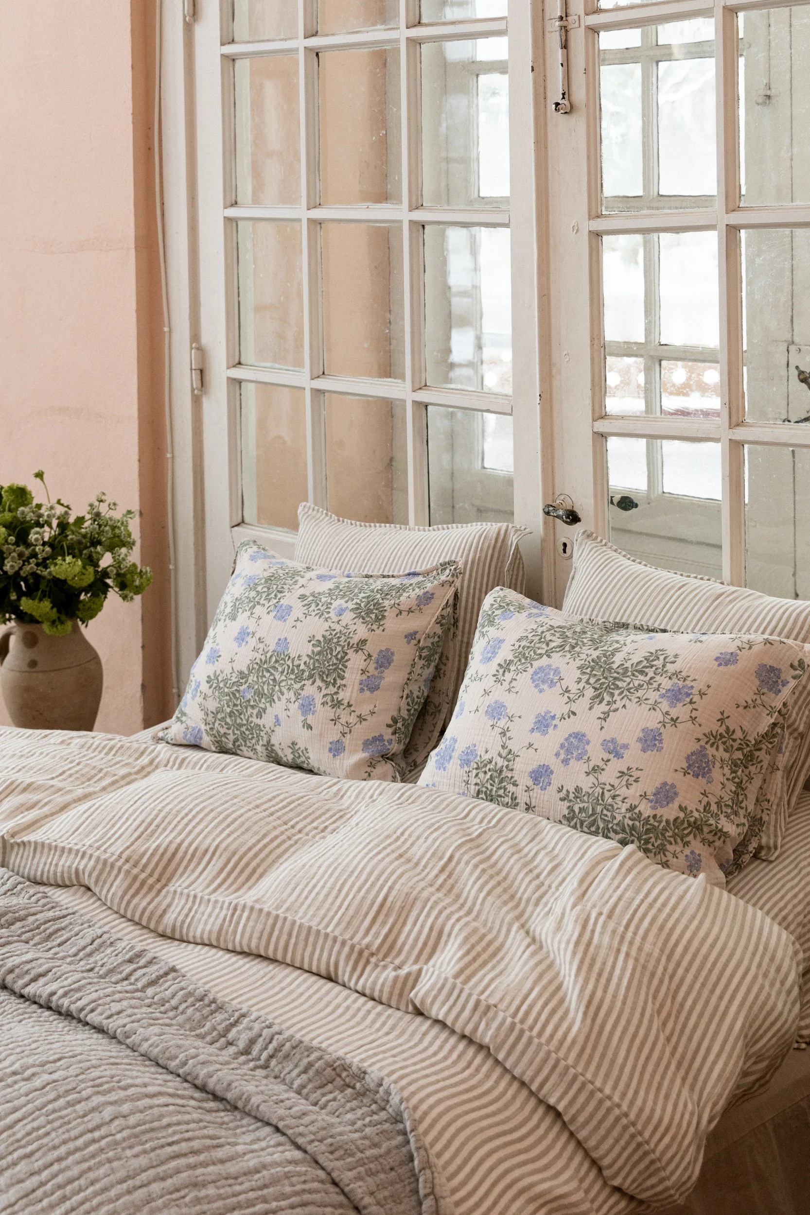 A bed with a striped duvet and two pillows with blue floral patterns, set against a paneled window. A vase with greenery is on the left.