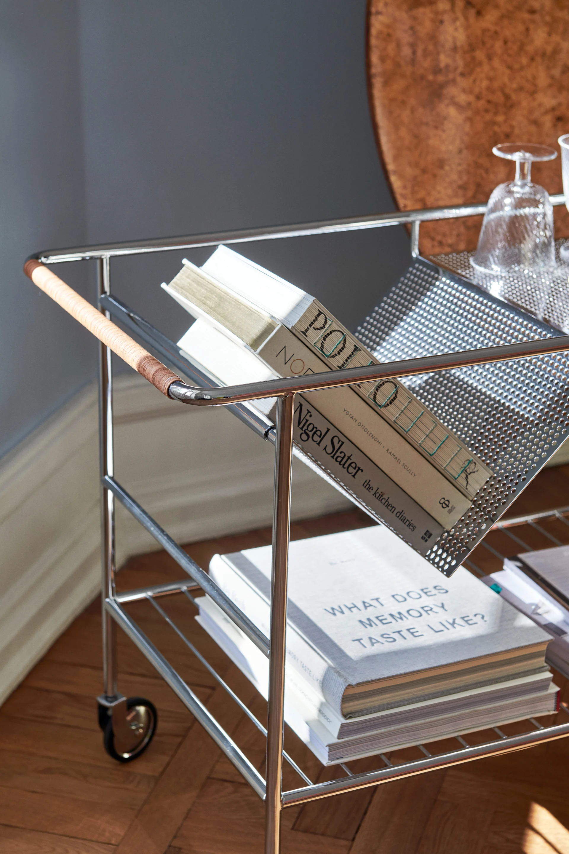 Chrome bar cart with a woven handle, holding books ('POLPO', 'WHAT DOES MEMORY TASTE LIKE?') and glassware.