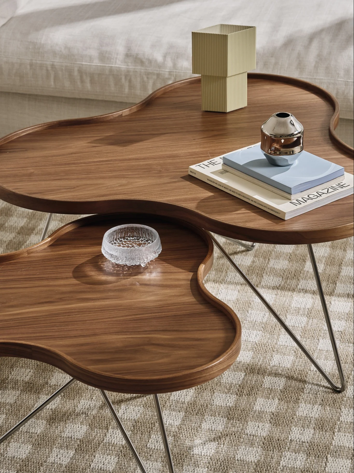 Two organic-shaped dark wood coffee tables, one holding a ribbed square object, metallic vase, and magazines, the other a glass bowl.
